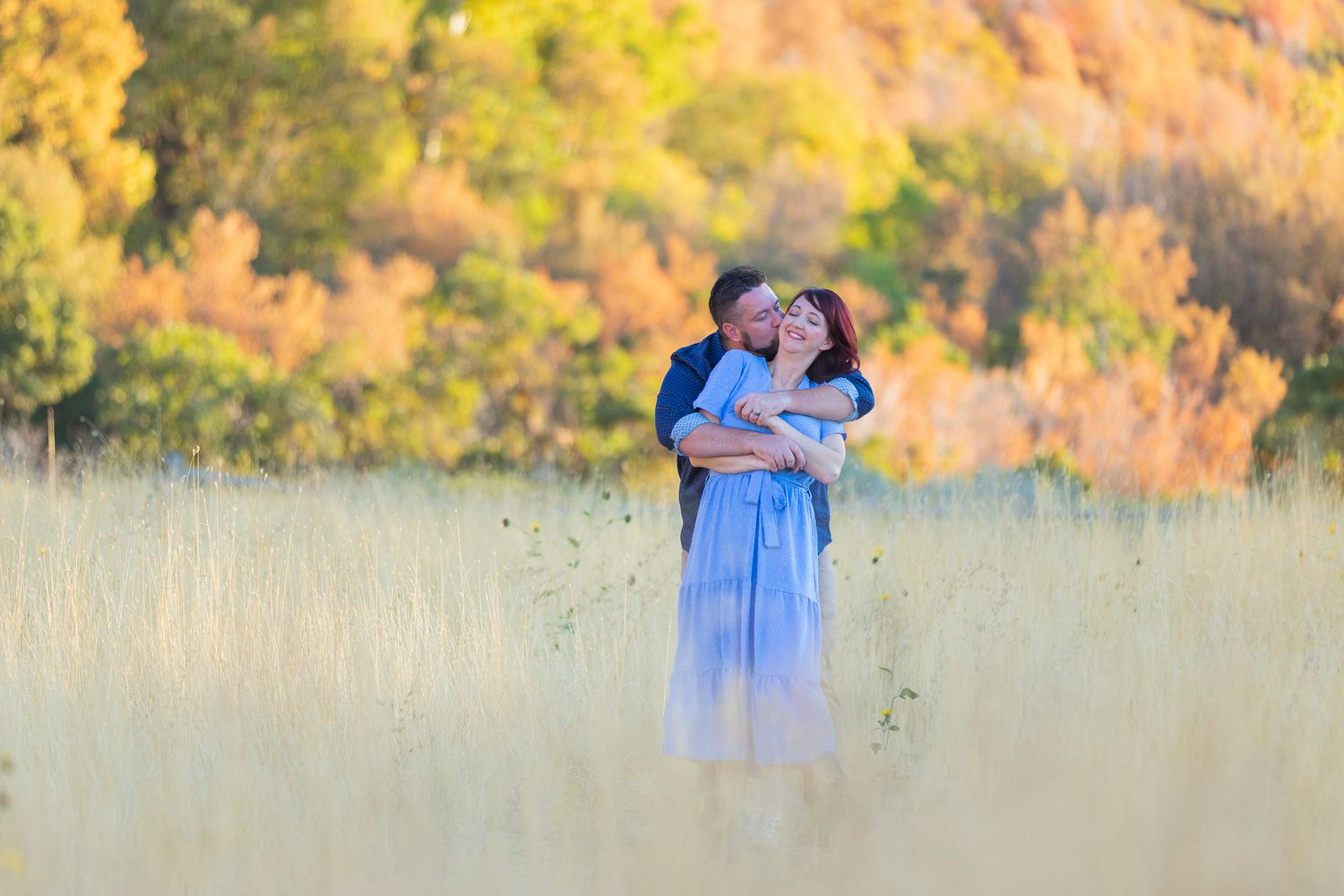 Couple embraces in tall grass field with golden autumn trees in background during sunset in Sandy, UT.