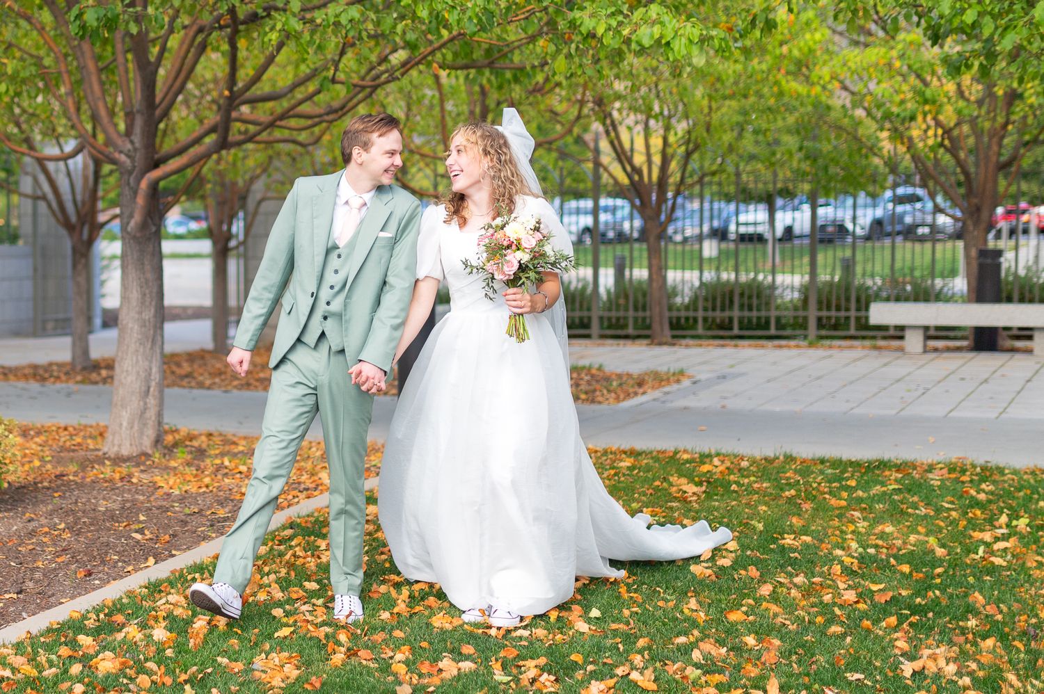 Happy newlyweds laugh together on path with autumn leaves in an outdoor setting.