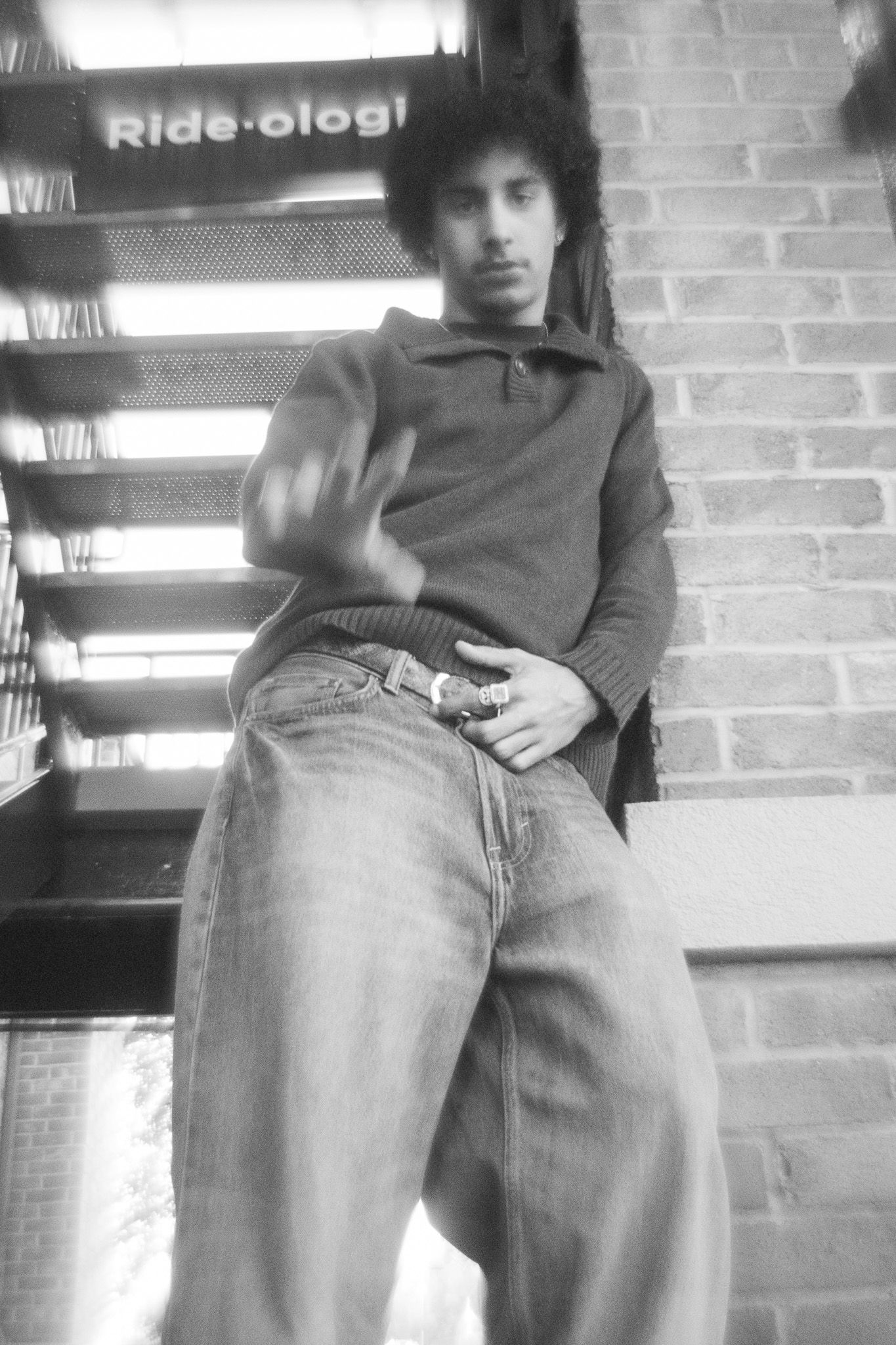 Black and white portrait of someone in casual attire leaning against brick wall with wide-leg jeans.