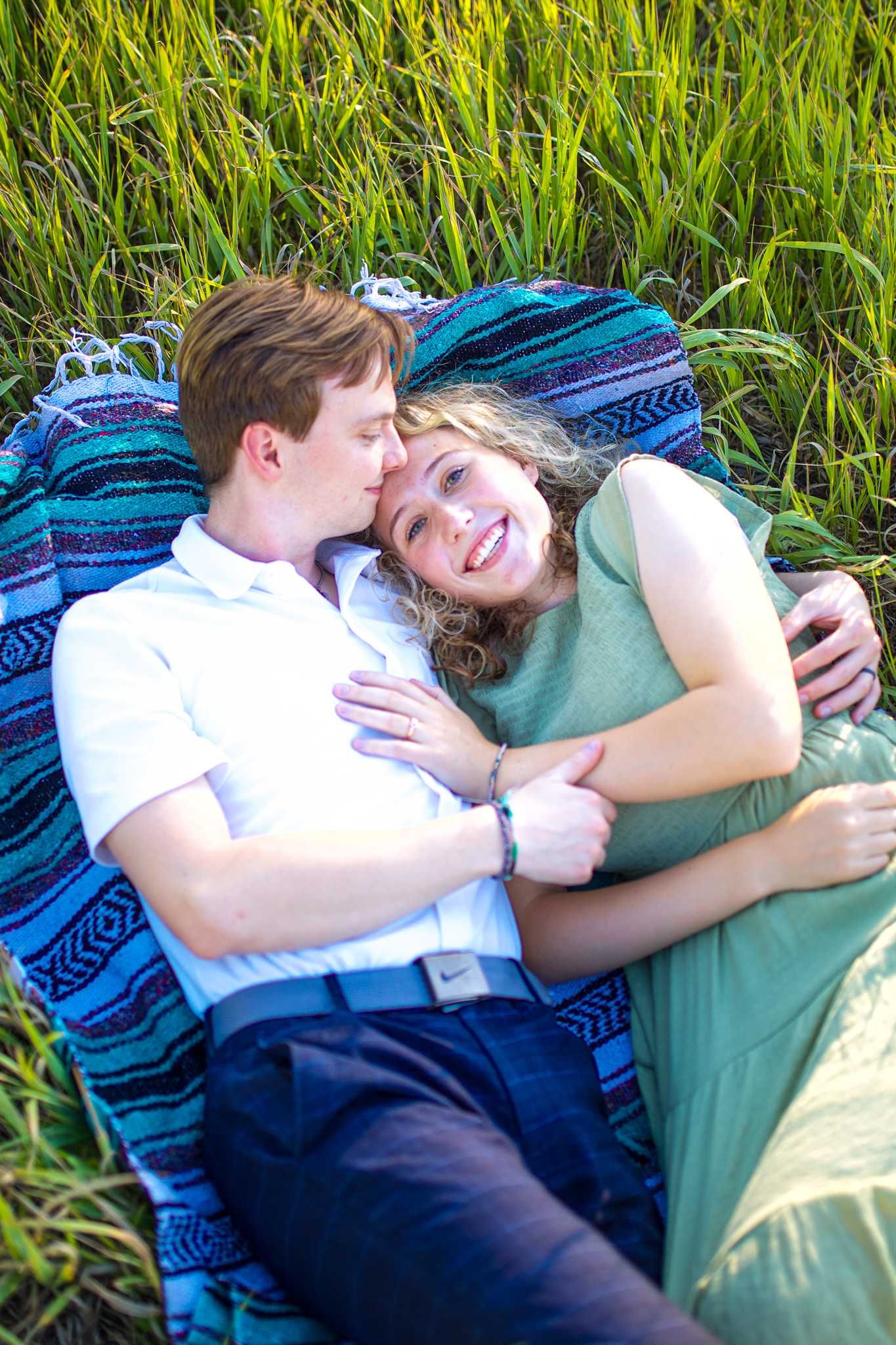A couple sharing an intimate moment while lying on a blue blanket surrounded by tall grass on a sunny day.