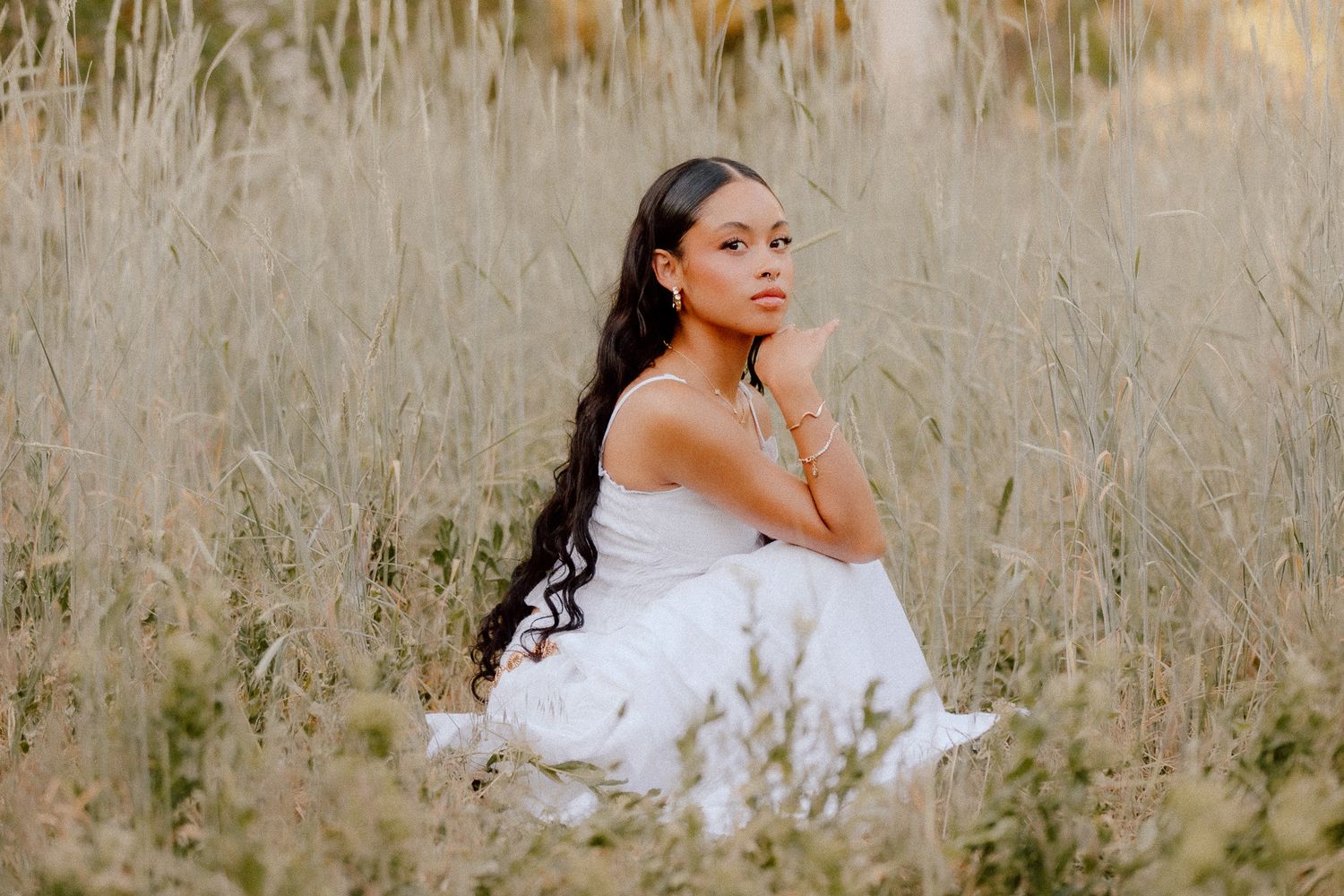 A person in a white flowing dress sits thoughtfully in a sunlit field of tall dried grasses in downtown Salt Lake City.