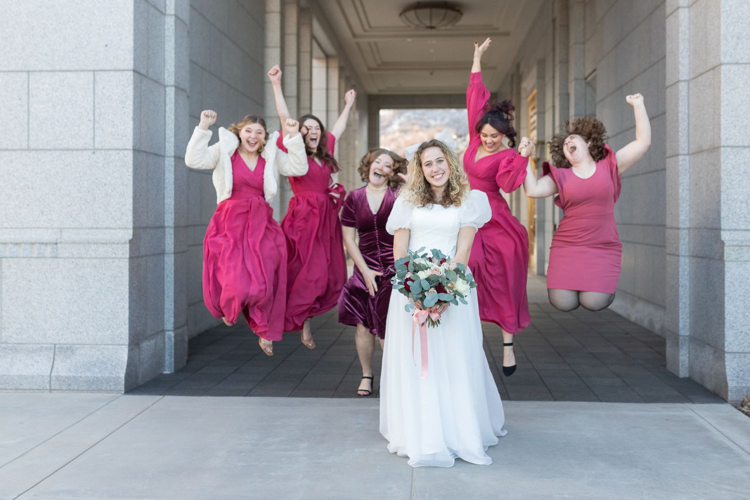 Wedding party jumps joyfully in archway with bride in white and bridesmaids in vibrant pink dresses.