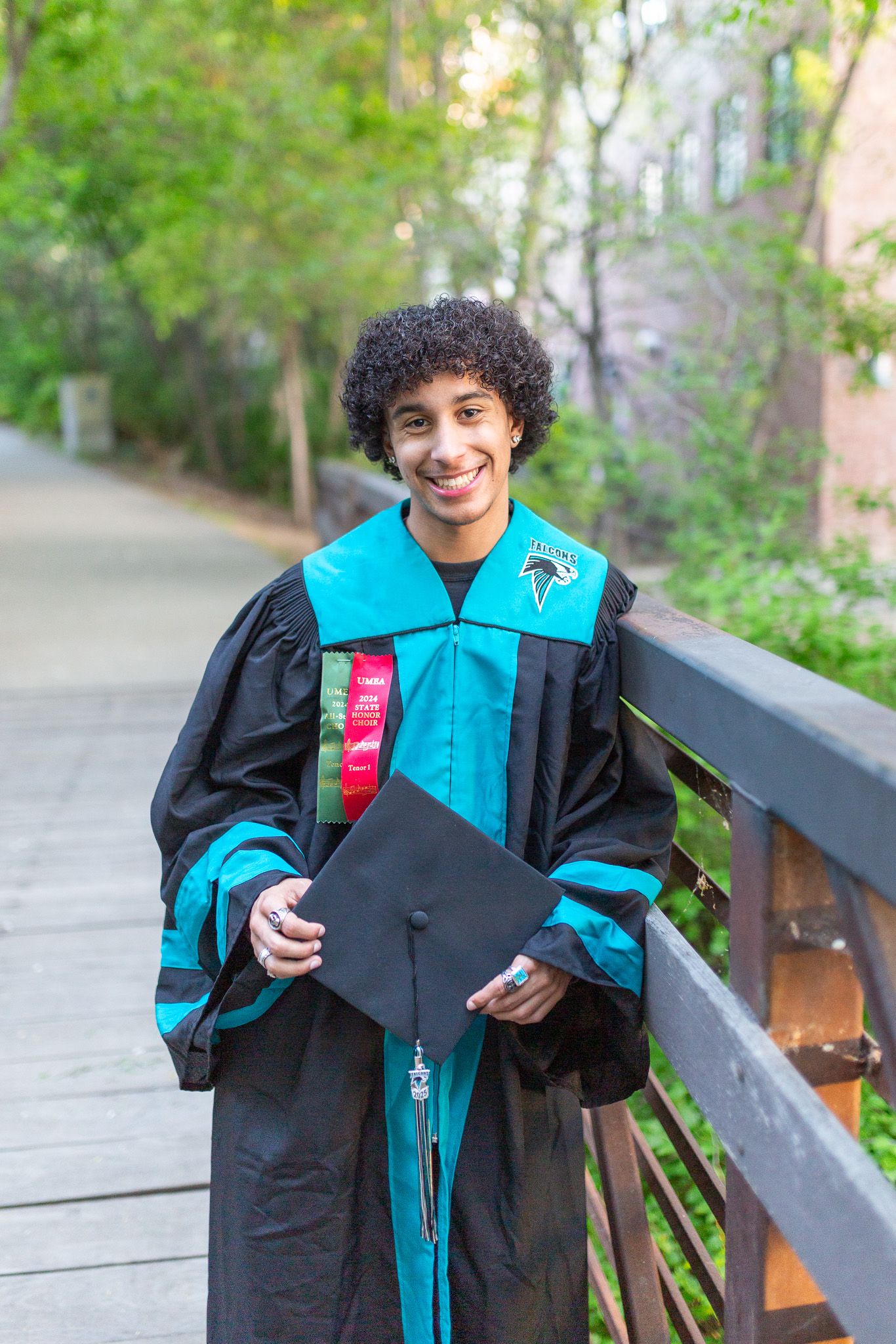 Graduate wearing teal and black graduation gown poses with diploma and beverage in outdoor setting.