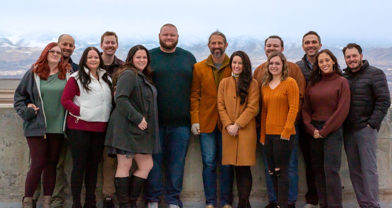 Group of people standing together outdoors wearing casual fall attire posing against mountain backdrop.