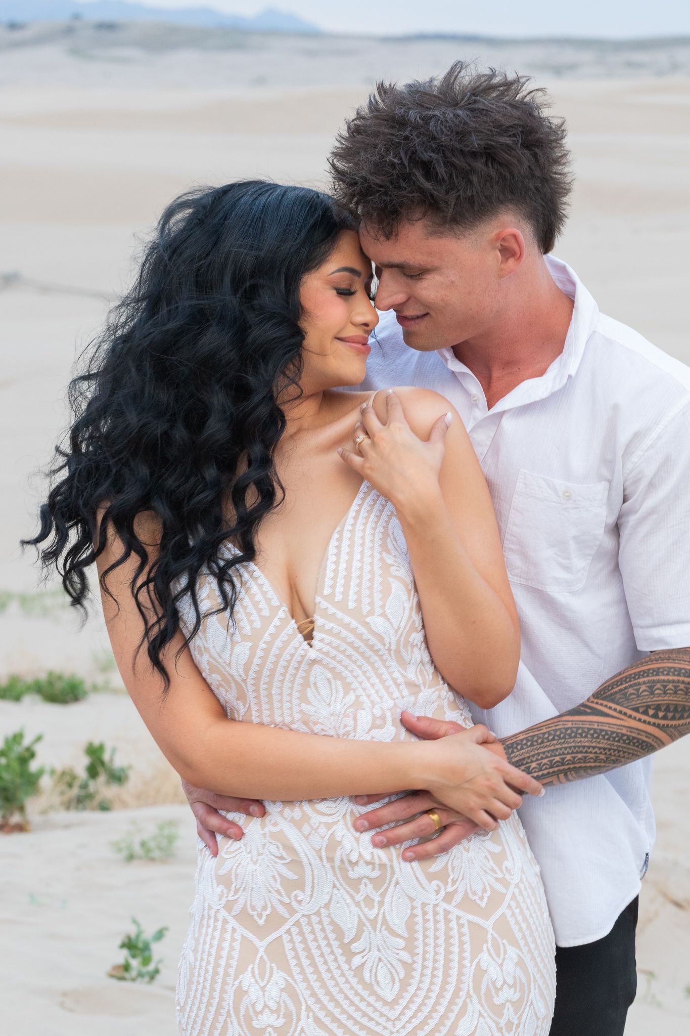 Couple embraces on sandy beach wearing white formal attire during romantic photoshoot.