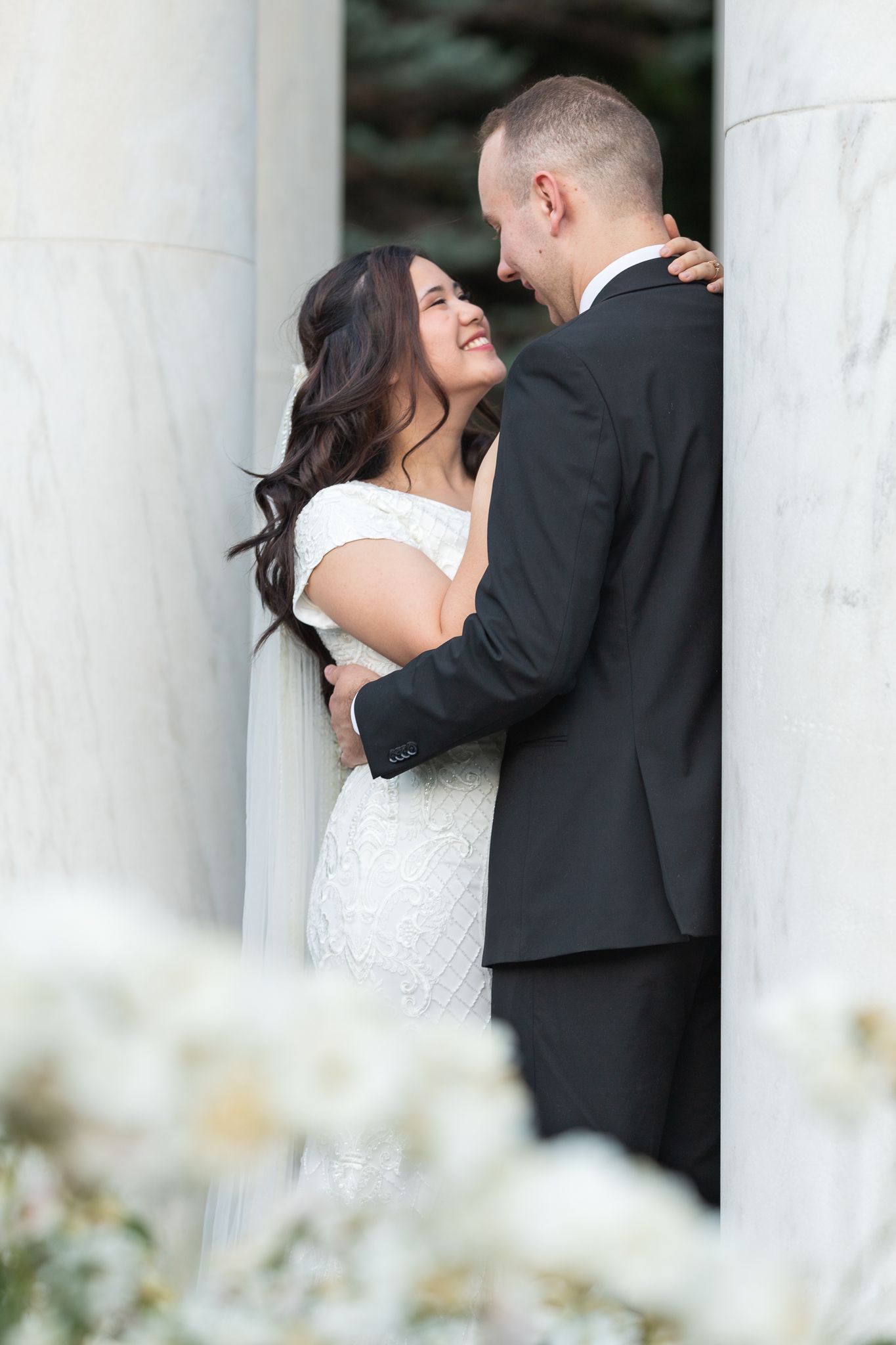 Couple shares intimate moment between white marble columns during formal photoshoot.
