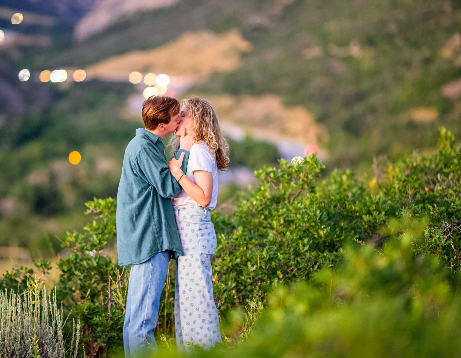 A romantic sunset moment shared between two people standing among green bushes with city lights glowing in background.