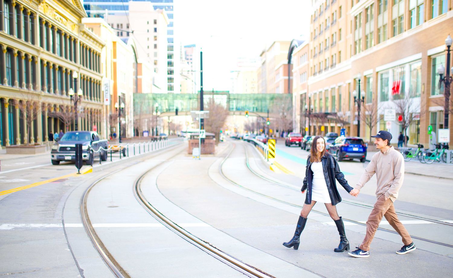 Couple walking across streetcar tracks in downtown area with tall buildings.