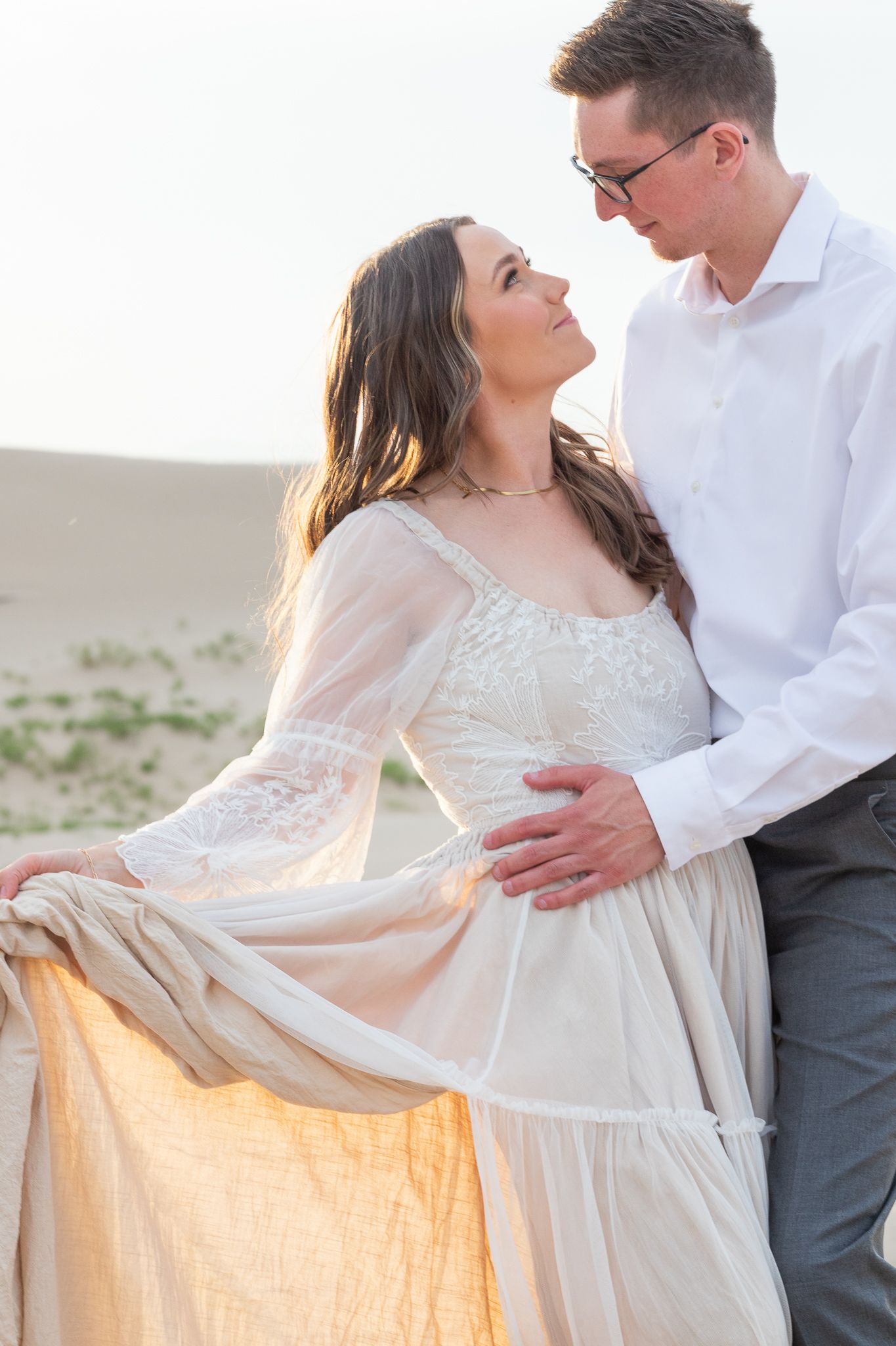 Romantic couple poses in white attire against a desert backdrop during dreamy sunset photoshoot.