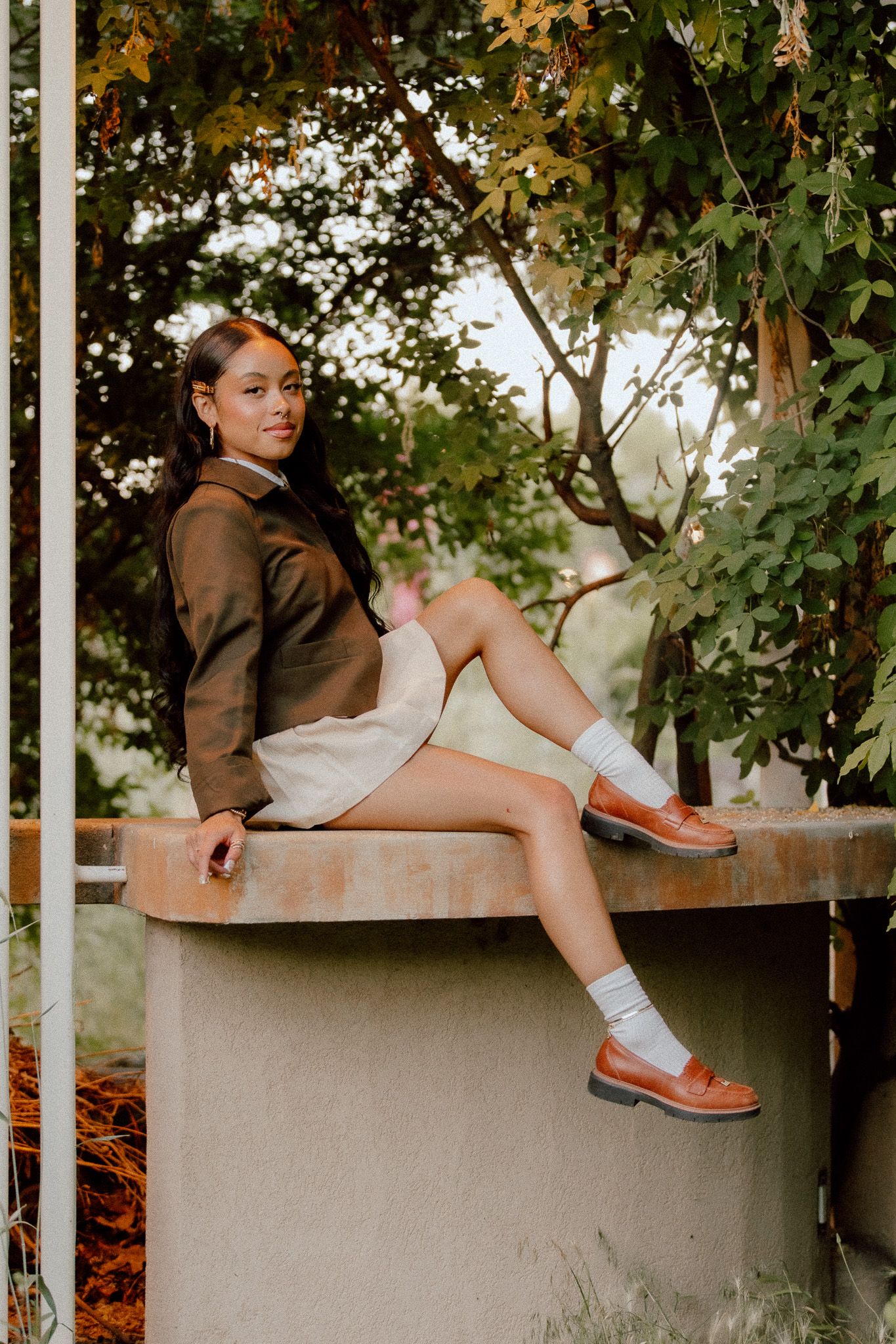 Woman in brown jacket and white dress reclines on concrete ledge surrounded by green leaves in a Salt Lake City Garden.