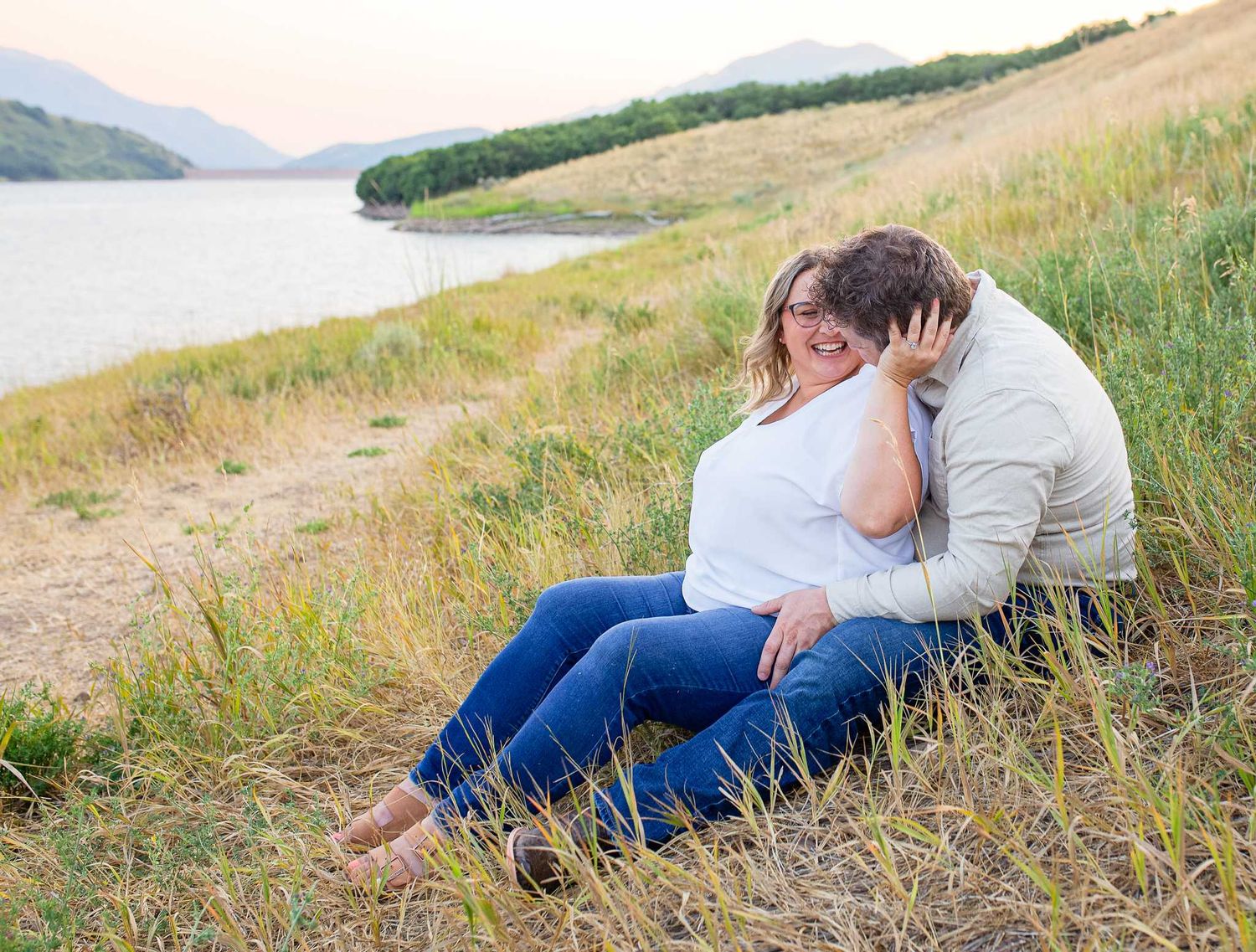 Couple sharing an intimate moment together on a grassy hillside overlooking a serene lake and mountains.