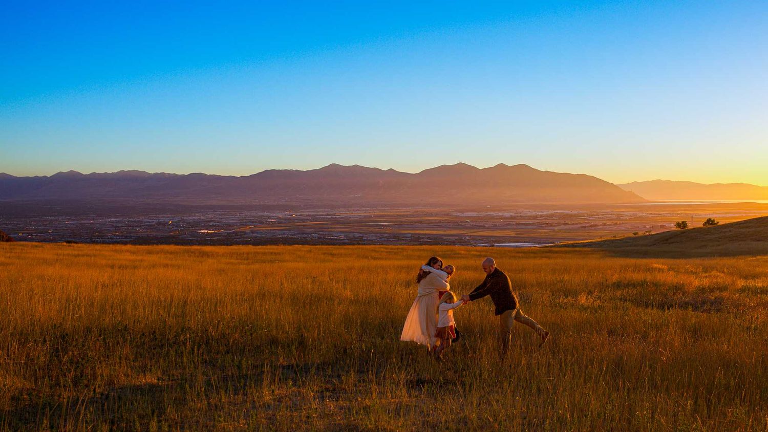 Two people sit together in a golden grass field watching the sunset over distant mountain silhouettes against a blue sky.