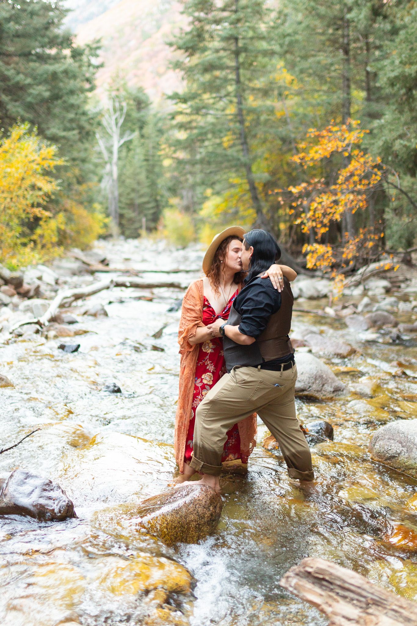 A couple shares a romantic moment by a mountain stream surrounded by autumn foliage and evergreen trees in Little Cottonwood Canyon, Utah.