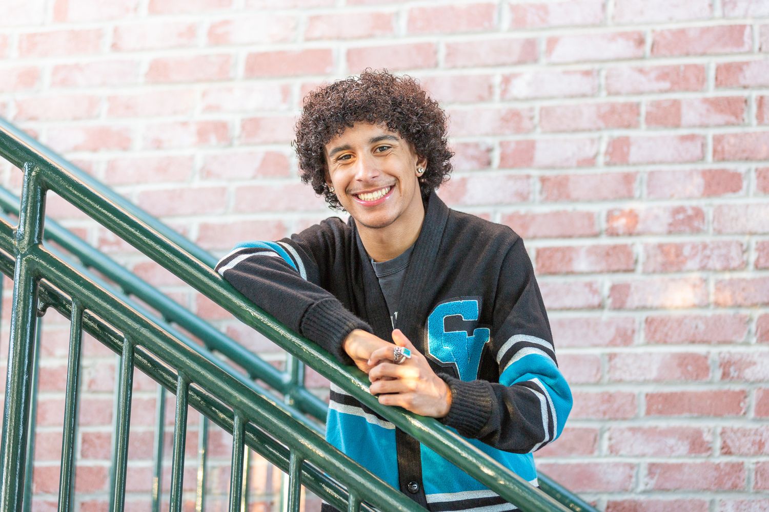 A student in a varsity letterman jacket leans casually on a metal railing against a brick wall background.