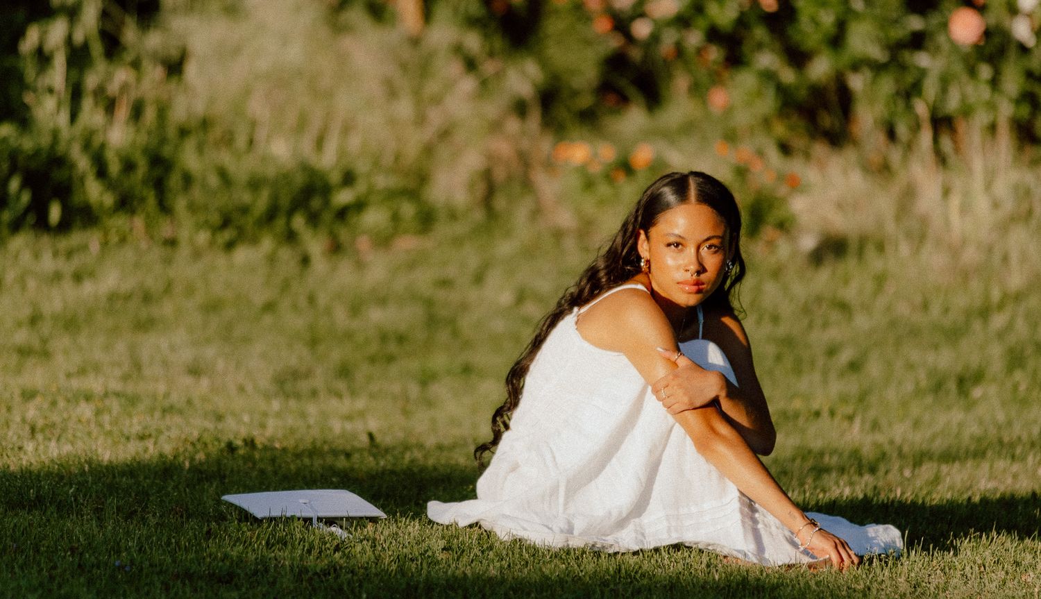 A Young woman in a white dress sits on a blanket in a sunny grass field in Utah during golden hour.