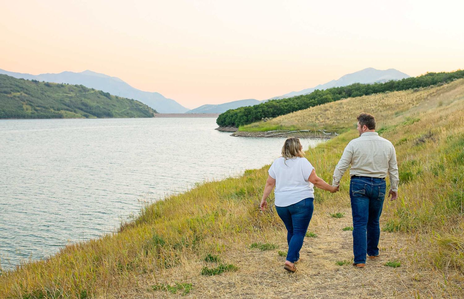 Couple walking hand in hand along a Utah lakeside trail with mountain views in the background.