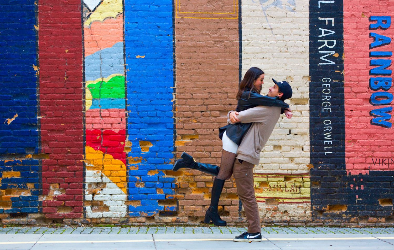 Couple embracing in front of vibrant colorful painted book spines mural on brick wall.