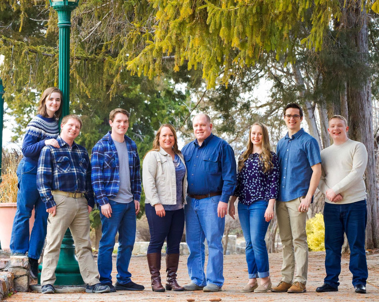 A group in coordinated blue and neutral tones stands together outdoors in front of evergreen trees during autumn.