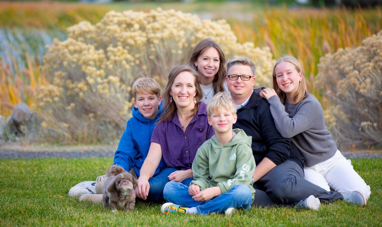 A group poses together outdoors on grass with autumn foliage in the background on a sunny day.