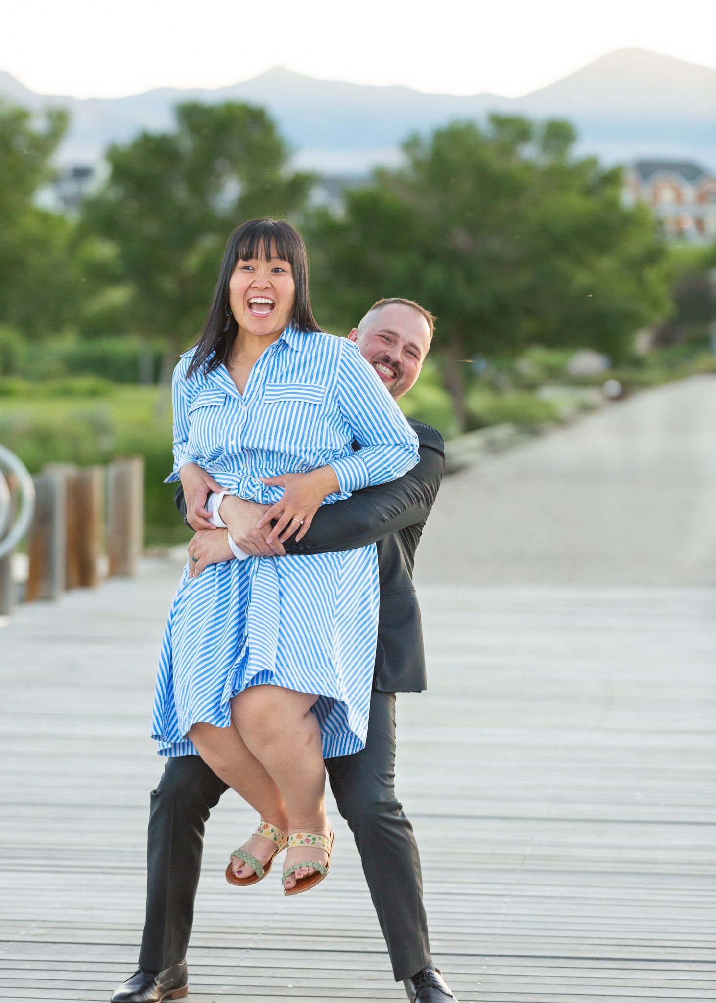 Playful couple sharing a joyful moment on a wooden boardwalk with mountains in the background.