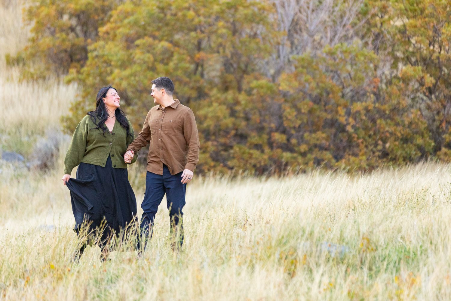 Couple walking through golden autumn field in Provo, Utah with fall foliage and trees in background during sunset.