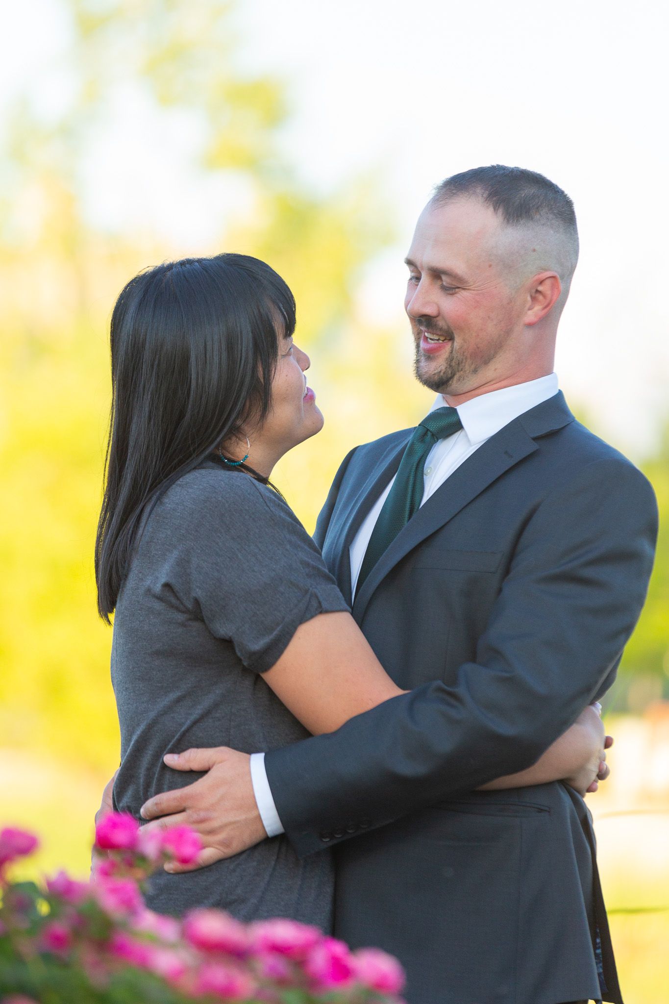 Romantic couple embraces during sunset engagement photoshoot with pink flowers in foreground.