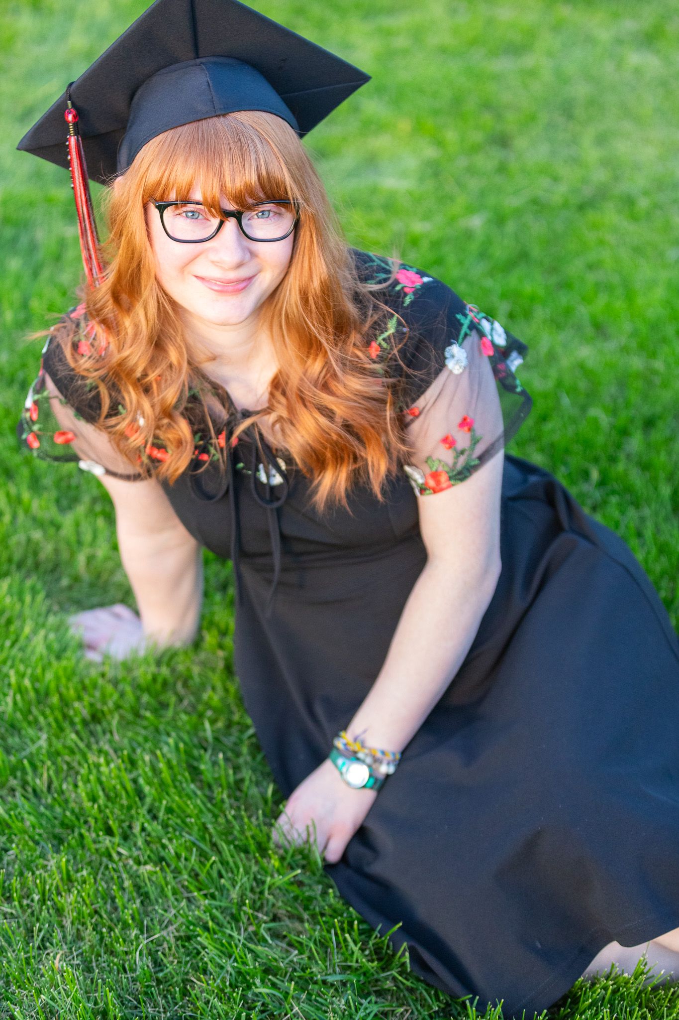 Graduate in black cap and gown with glasses sits on green grass during outdoor graduation photoshoot.