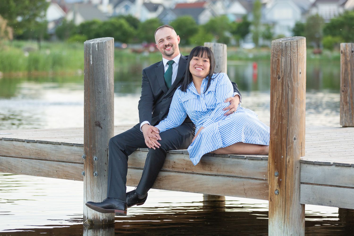 Couple sitting together on a wooden dock by the water with residential buildings in the background.