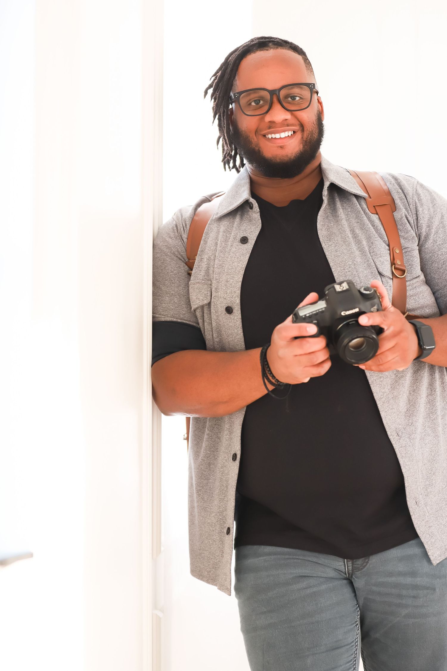Happy photographer wearing glasses and gray button-up shirt holds professional camera while smiling indoors.