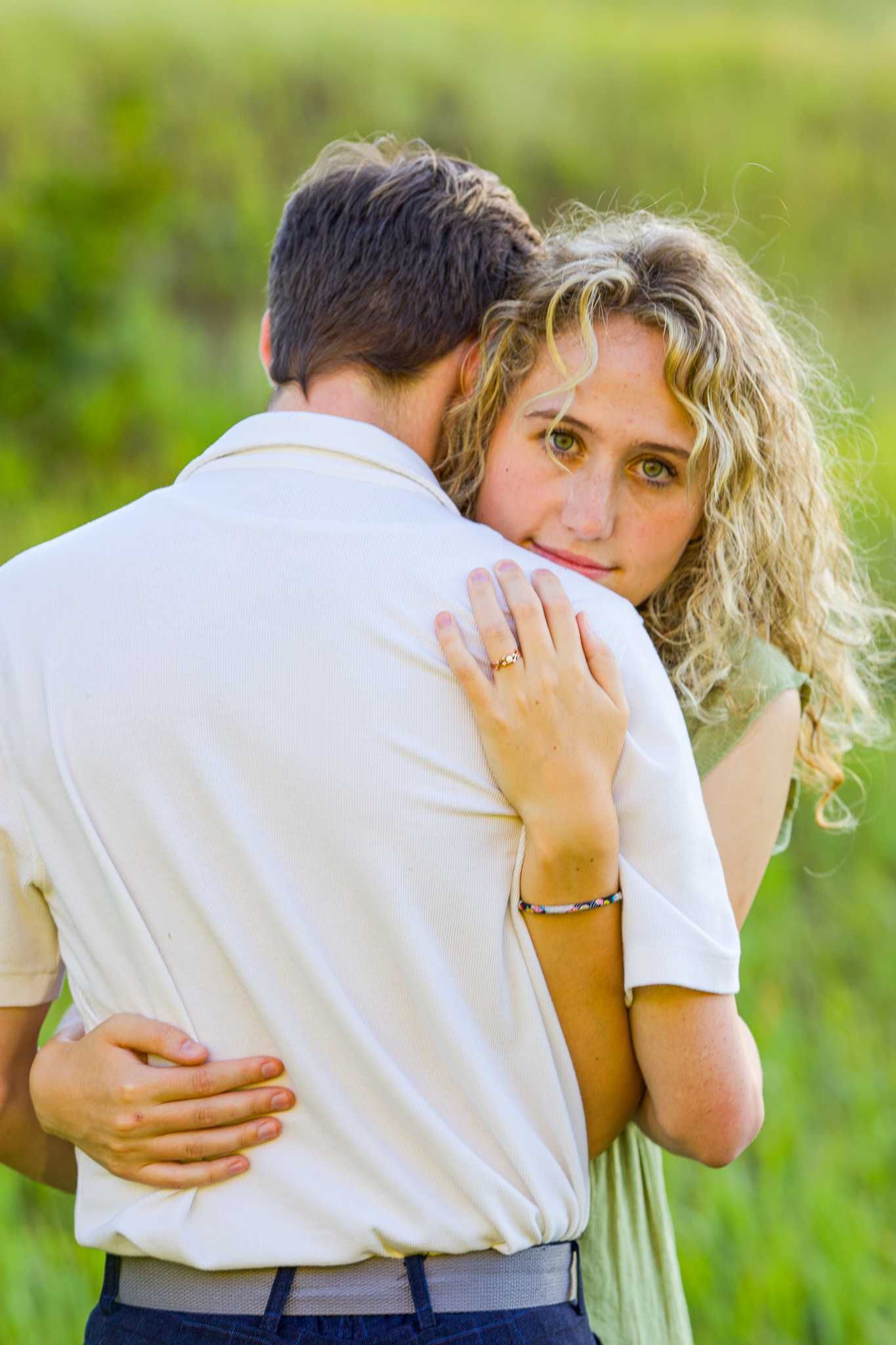 Engaged couple embraces in a sunny field while showing off the engagement ring.