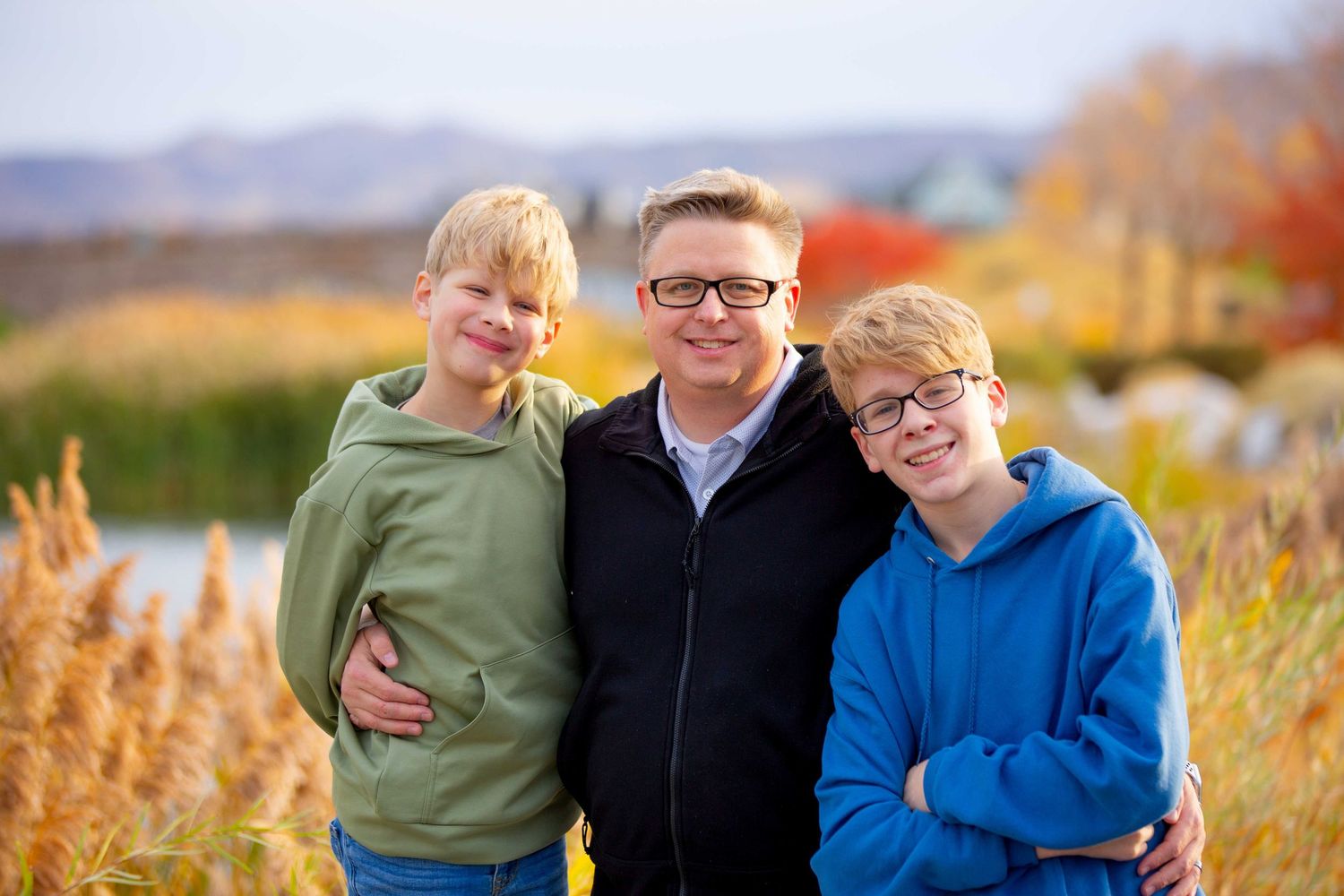 Three people in hooded sweatshirts stand together smiling outdoors on an autumn day with tall grass in the background.