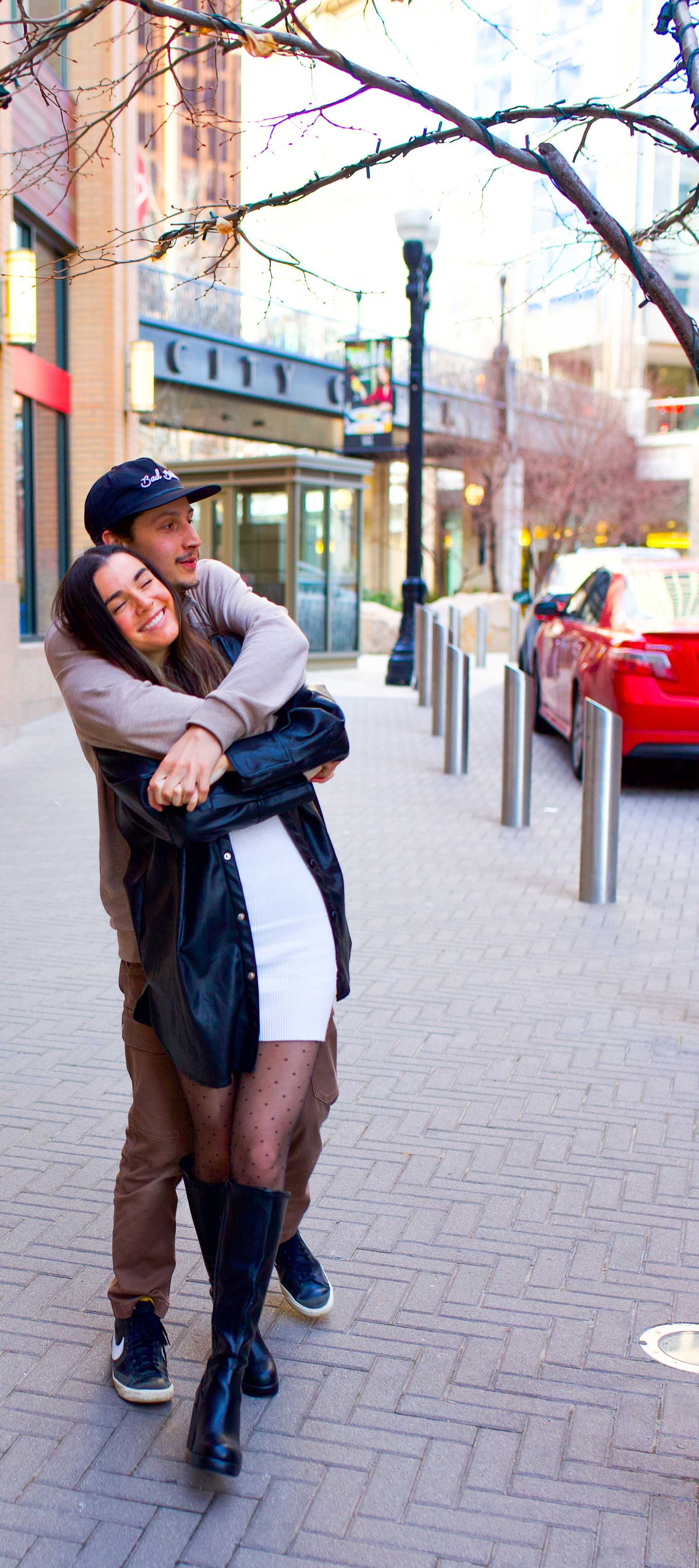 Couple sharing an intimate embrace on urban sidewalk with tree branches overhead.