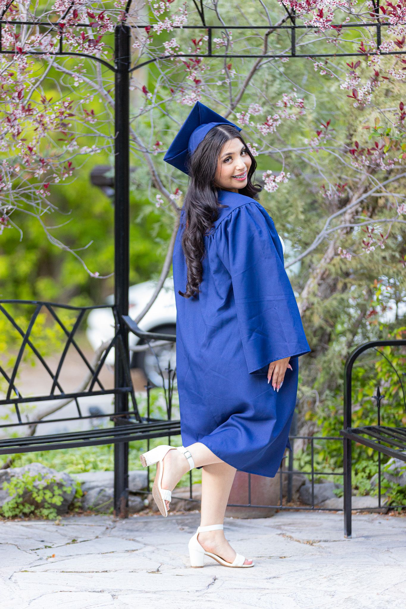 Graduate in blue cap and gown poses playfully by garden gate with pink blossoms in Millcreek Canyon, UT.