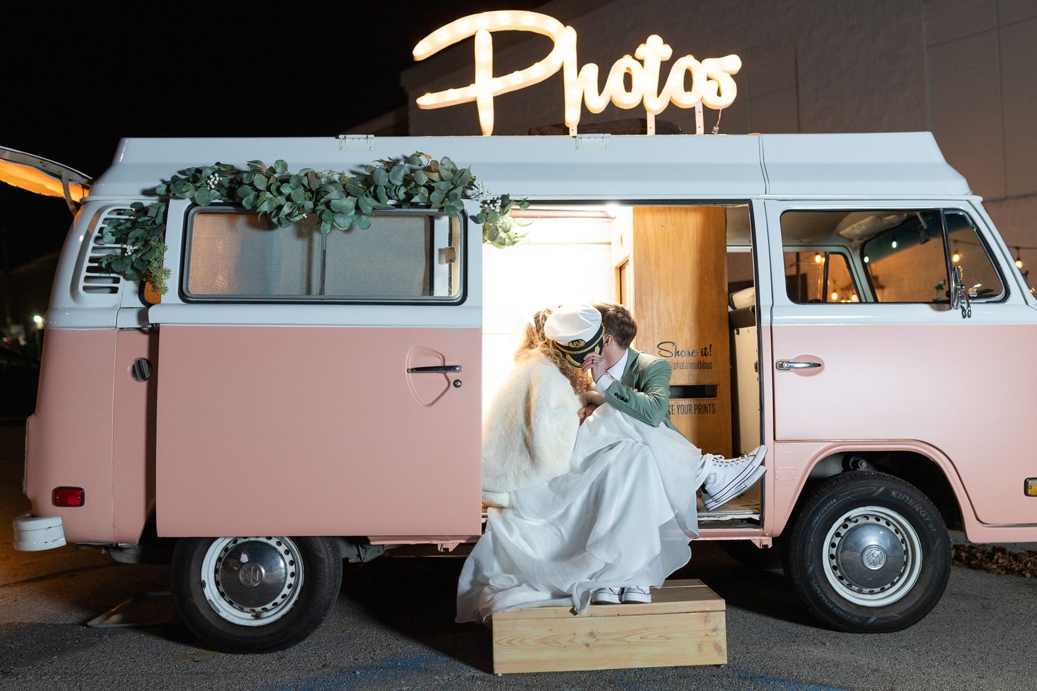 Wedding couple sitting in a vintage pink VW van with a neon 'Photos' sign above.