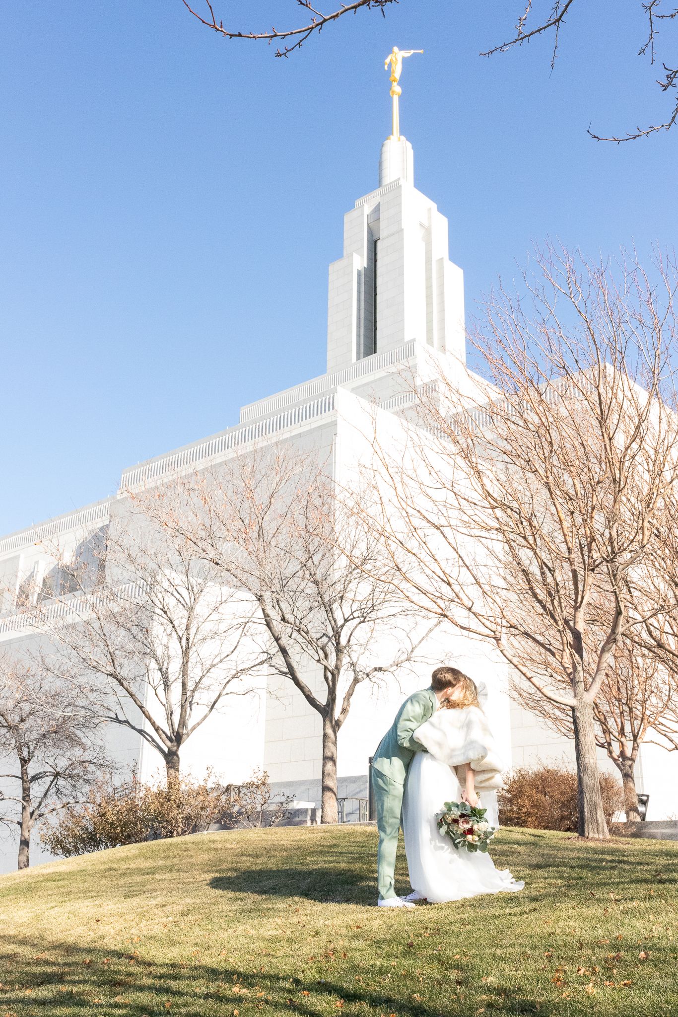 Wedding photo shoot near white church building with bare trees against blue sky.