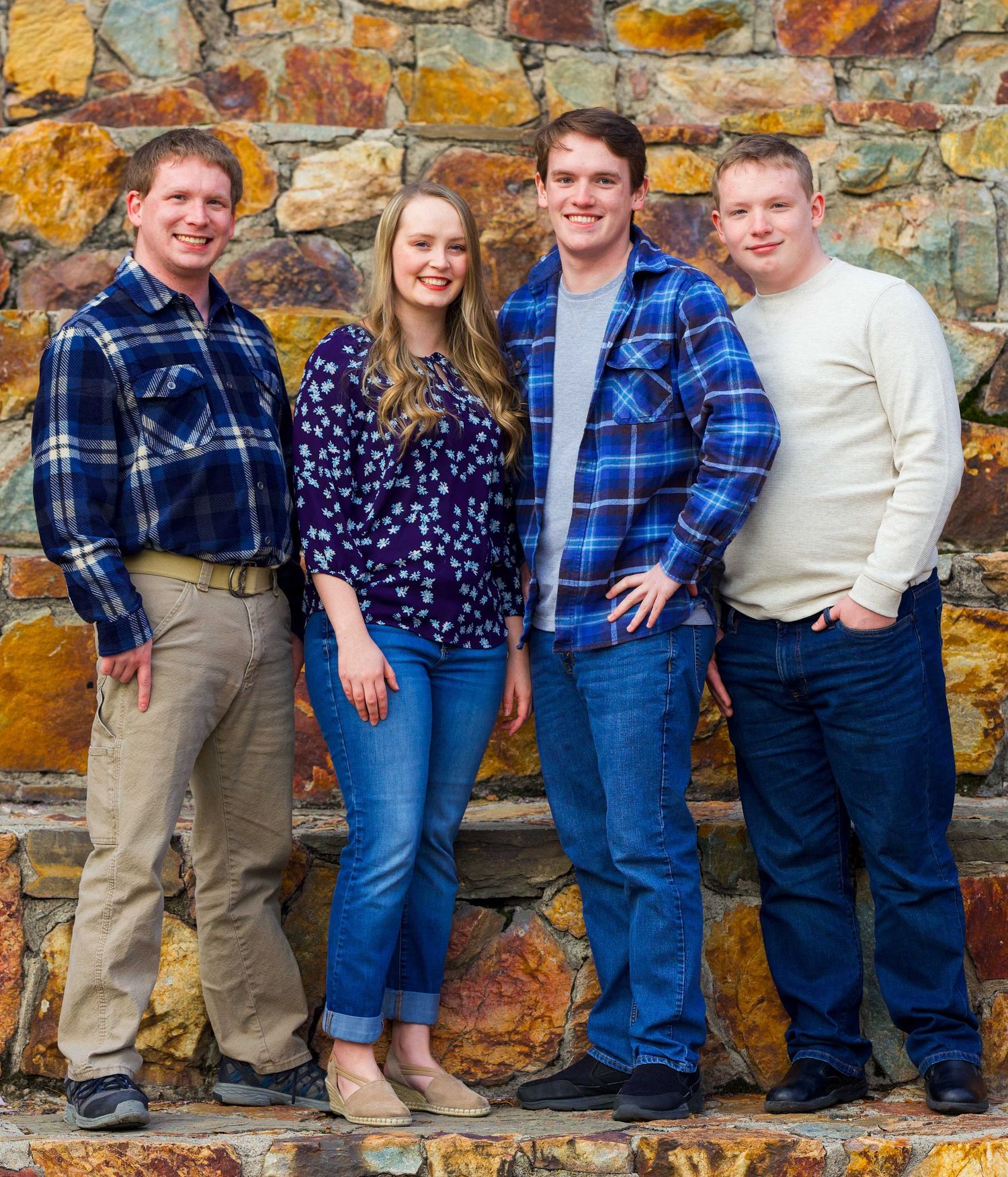 Four people stand together against a rustic stone wall, wearing casual clothing including plaid shirts and jeans.