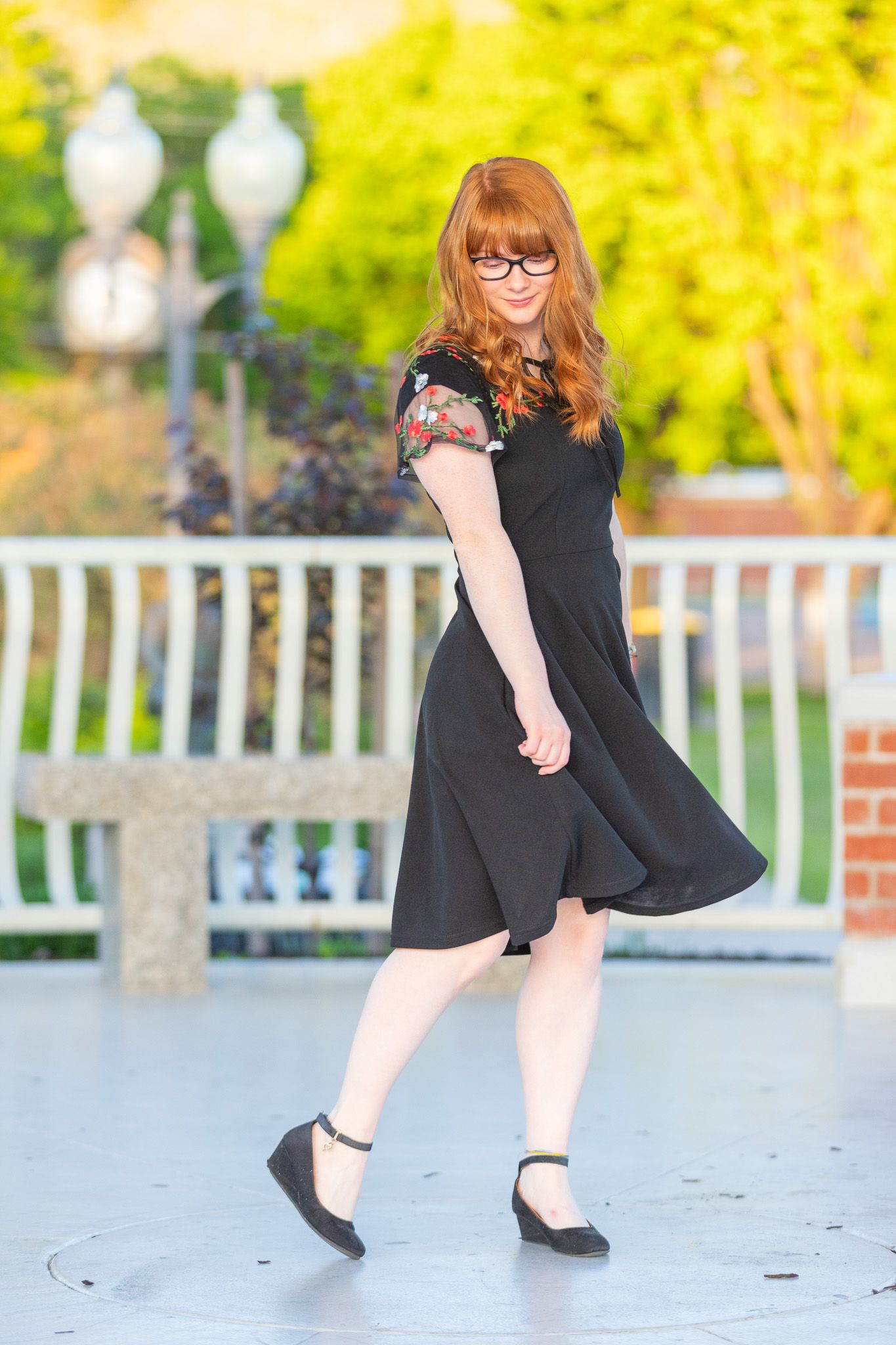 A woman in a black cap sleeve dress and flats poses on a porch with a decorative railing and yellow foliage in background.