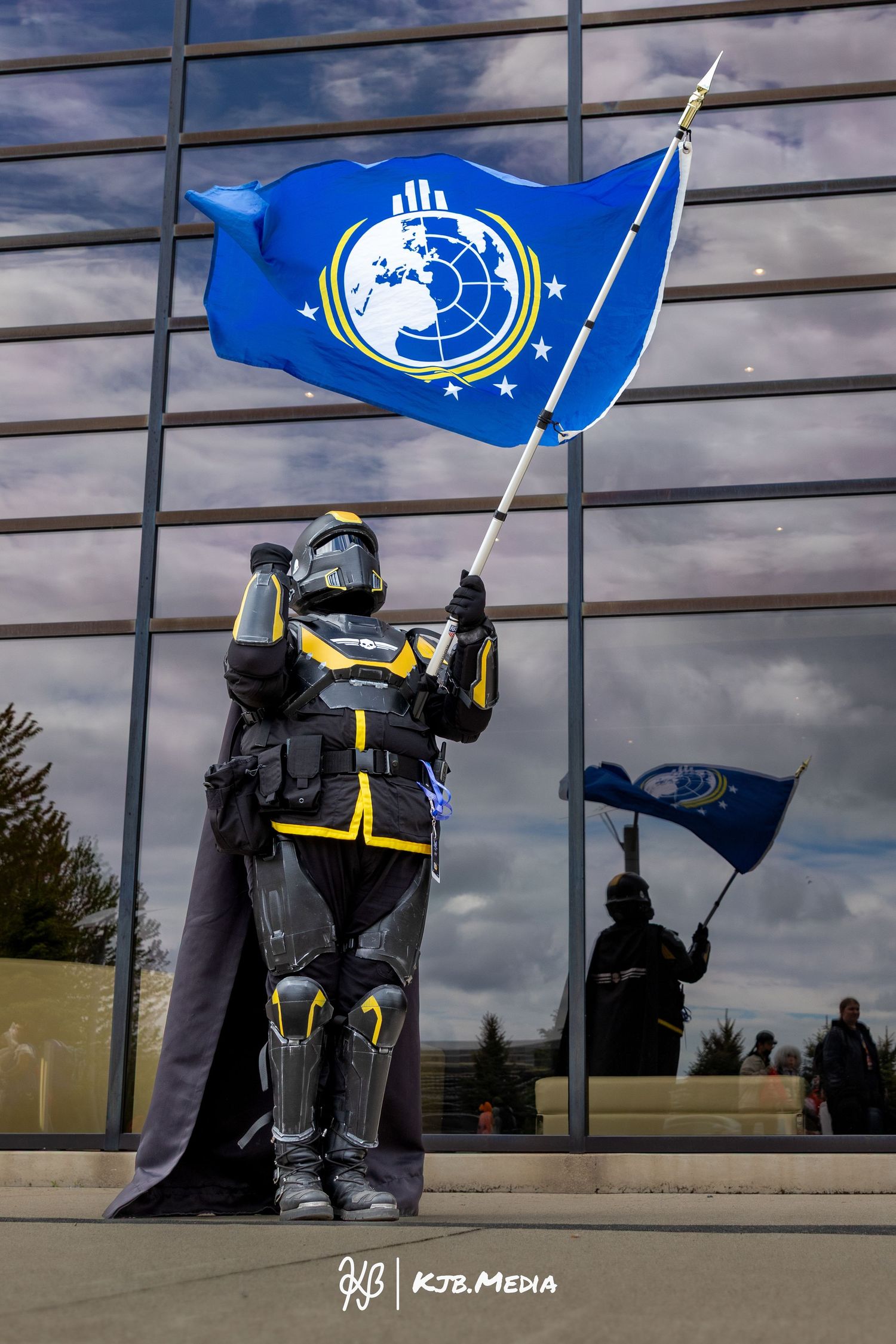 Costumed figure in armor holds a bright blue flag with an emblem against a modern building facade.