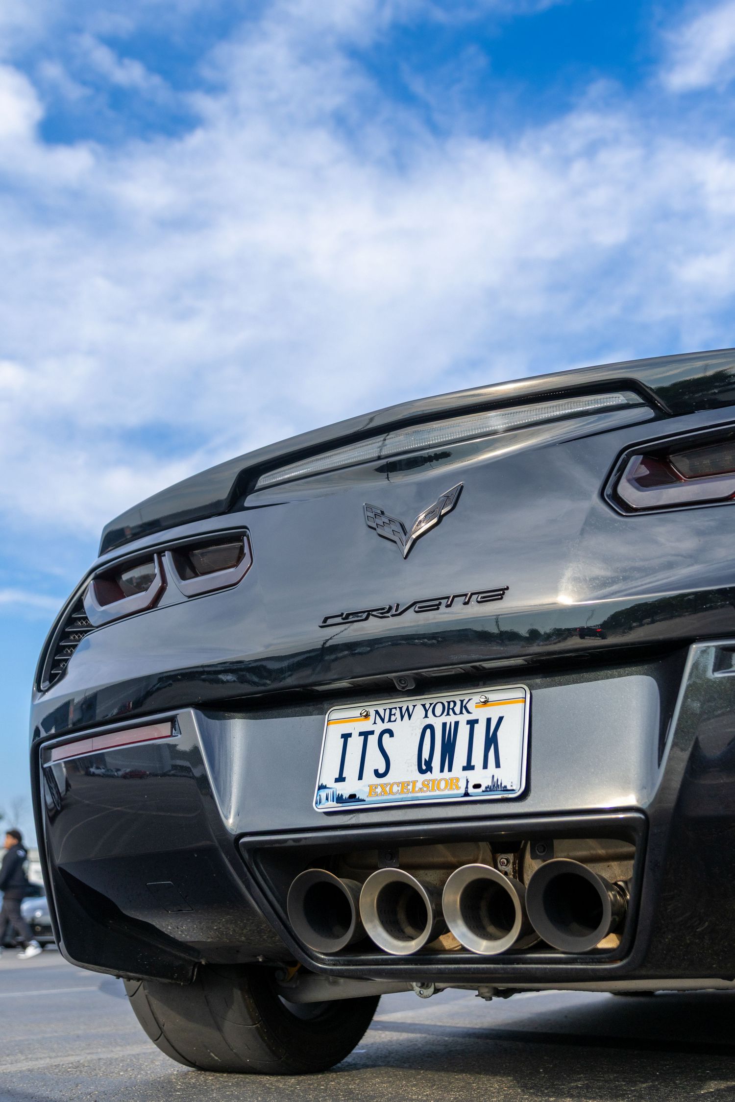 Low angle view of a black Chevrolet Corvette's rear end showing exhaust pipes and license plate against blue sky.