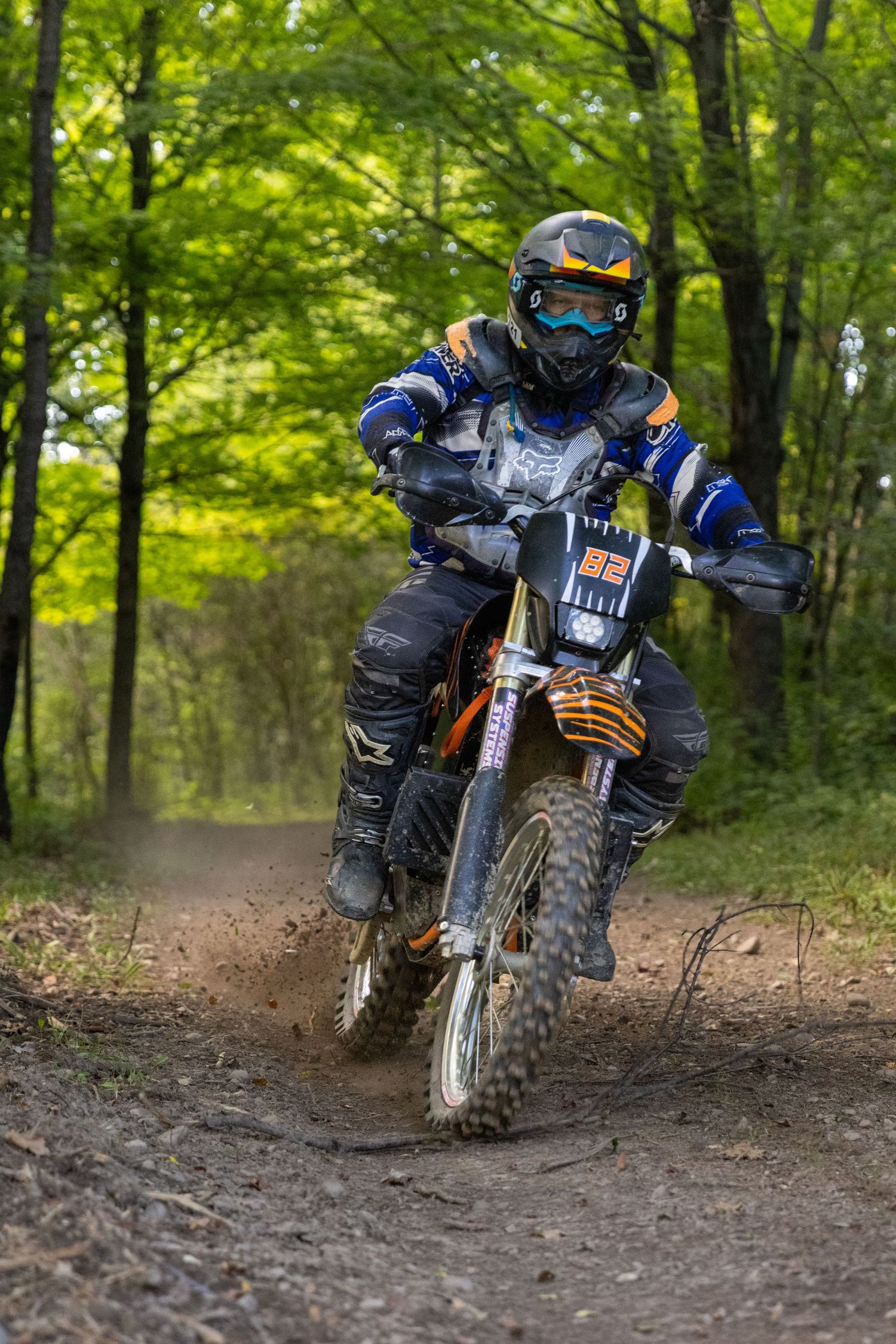 Motocross rider in blue gear navigates a dirt trail through a lush green forest on a muddy off-road motorcycle.