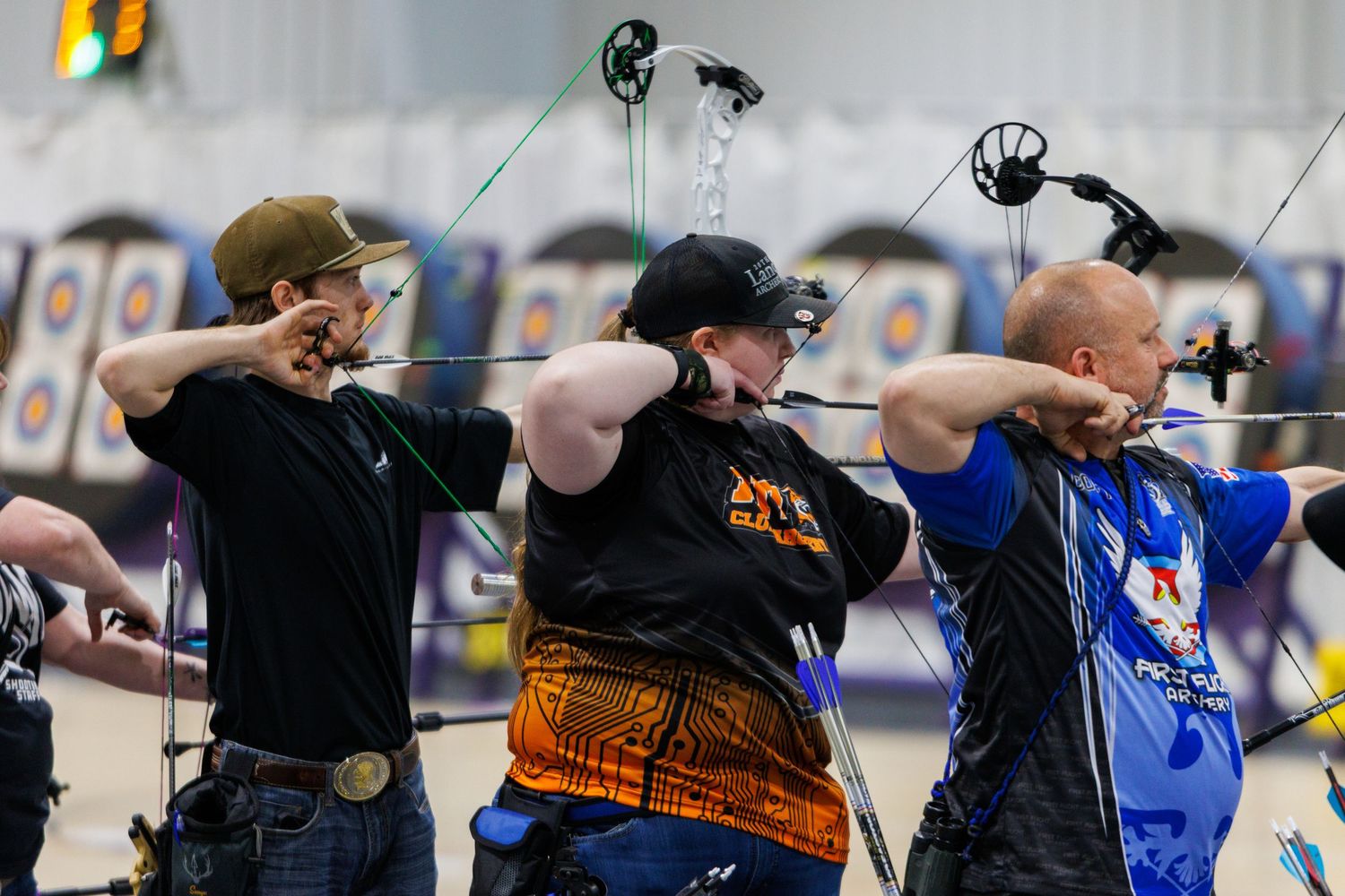 Three focused archers drawing their bows simultaneously during an indoor archery competition.