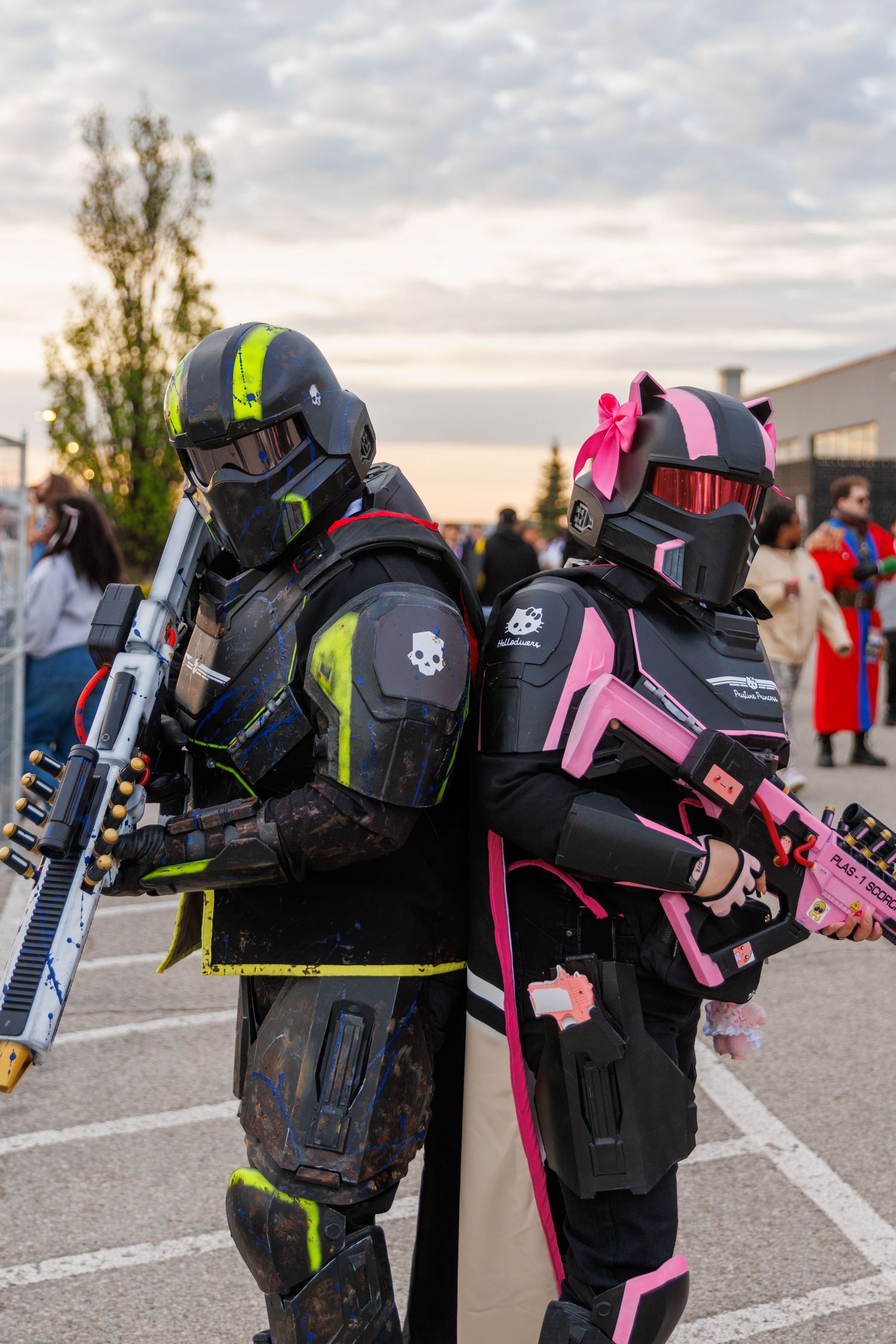 Two cosplayers in black and neon armor suits pose back-to-back holding prop guns at an outdoor convention.