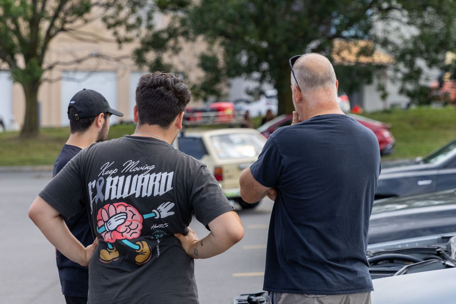People in black shirts gather around classic cars at an outdoor car show meet on a sunny day.
