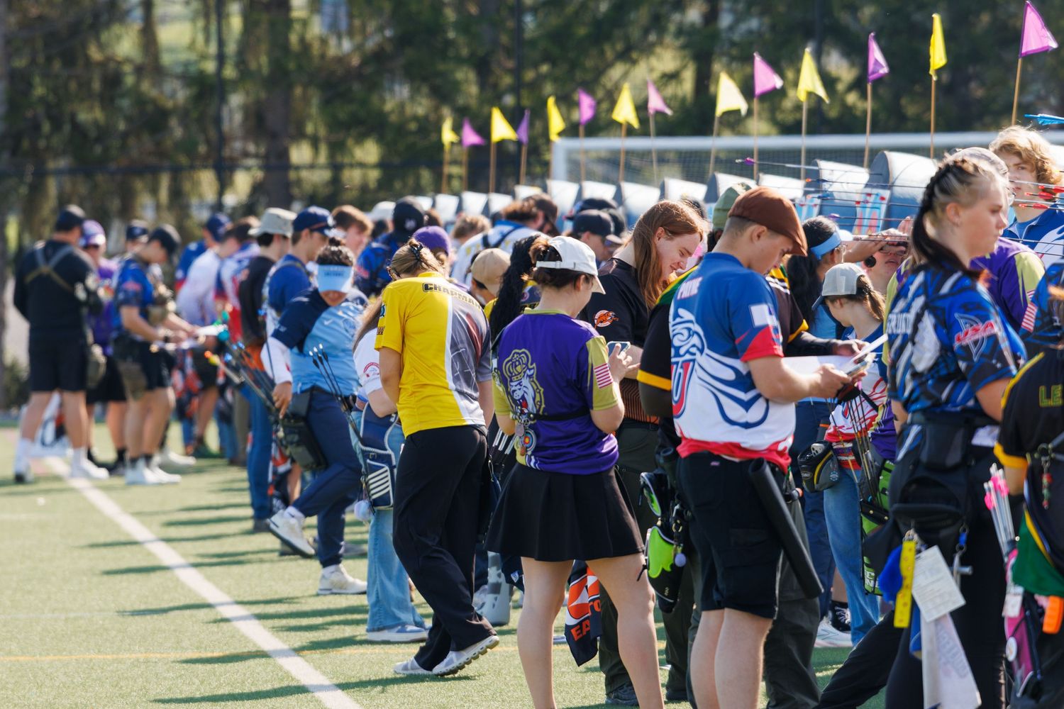 Archers line up on a green field at an outdoor archery competition, colorful flags visible in the background.