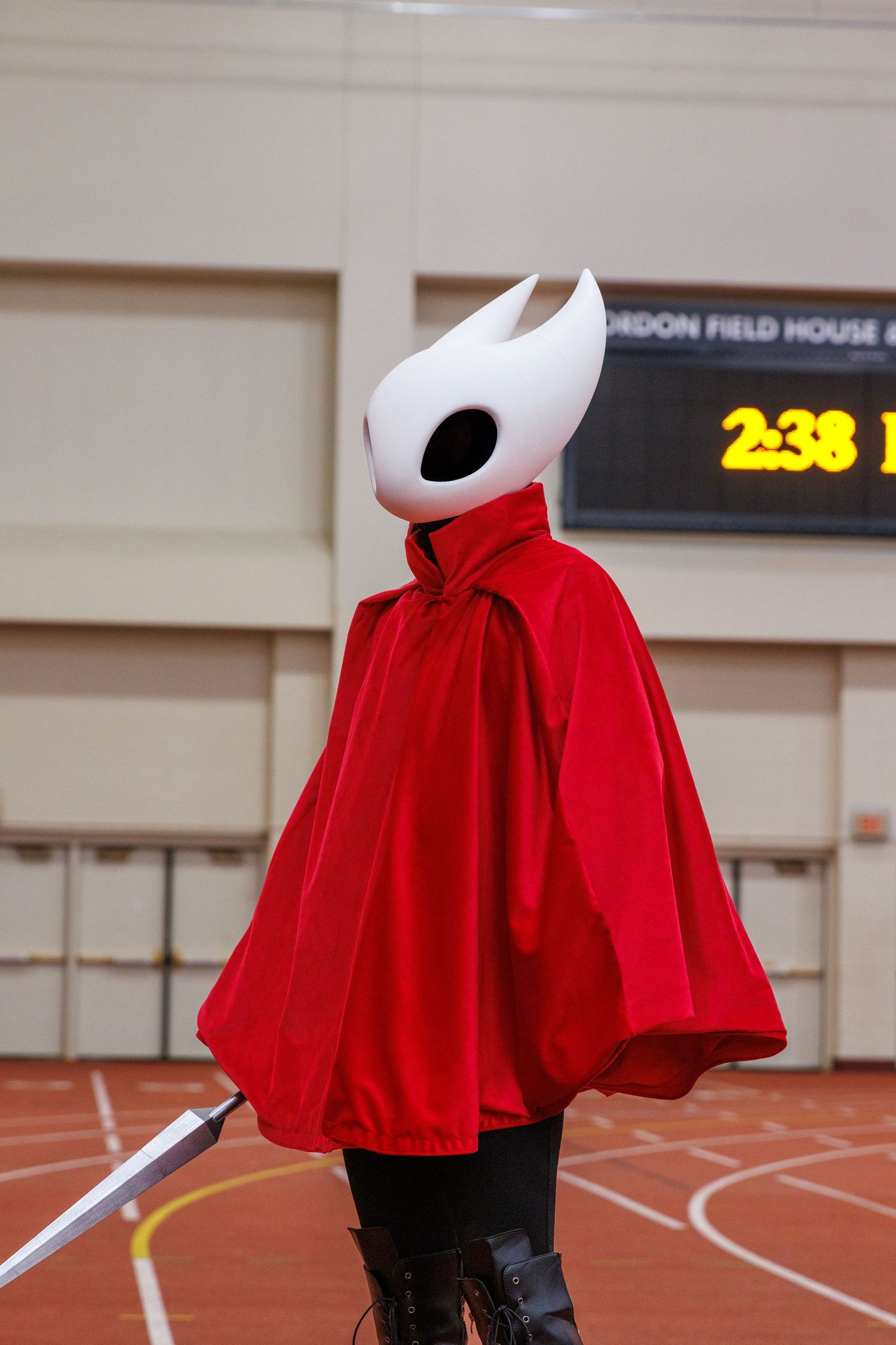 Person cosplaying as Hollow Knight's Hornet in red cape, white mask, and sword at an indoor track facility.