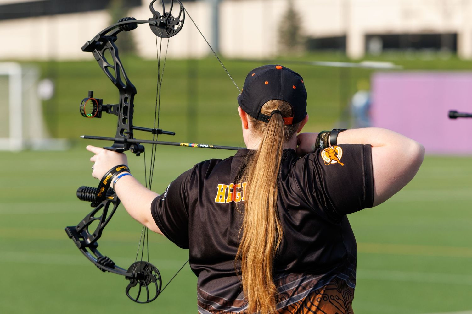 Female archer in black team shirt aims a compound bow on a green sports field during competition.