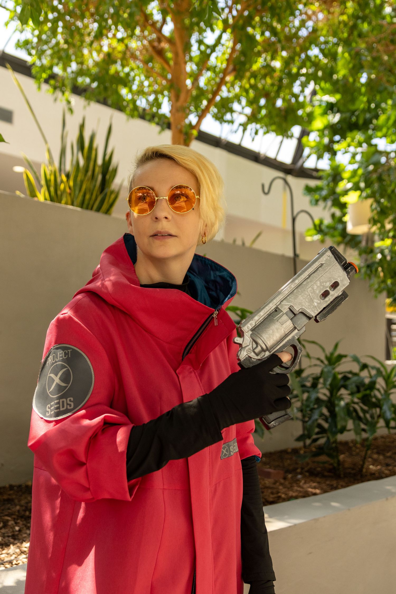 Person posing in a red hoodie and black gloves holding a prop weapon in front of greenery and white walls.