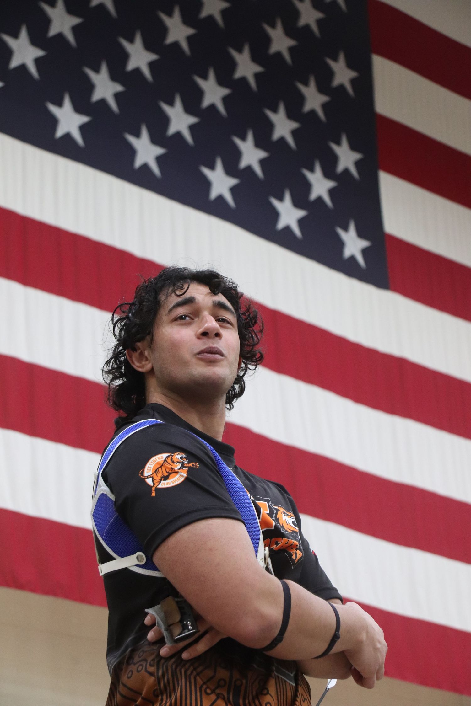 Person stands confidently in front of large American flag wearing archery equipment.