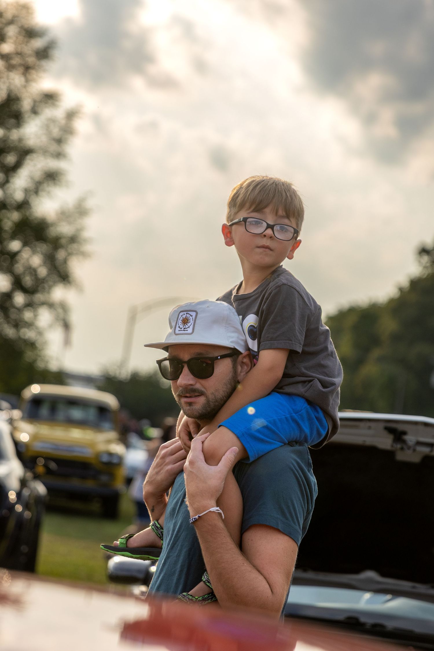 A parent and child share a tender moment outdoors on a cloudy day with parked cars visible in the background.