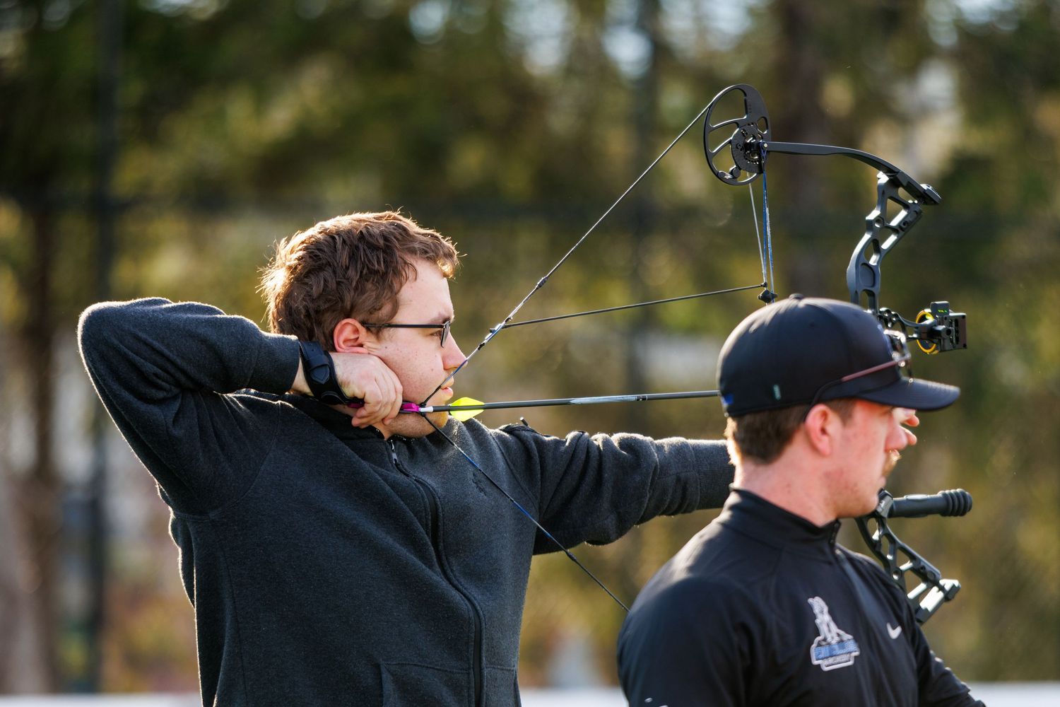 Young archer drawing a compound bow with a teammate standing nearby at an outdoor range.